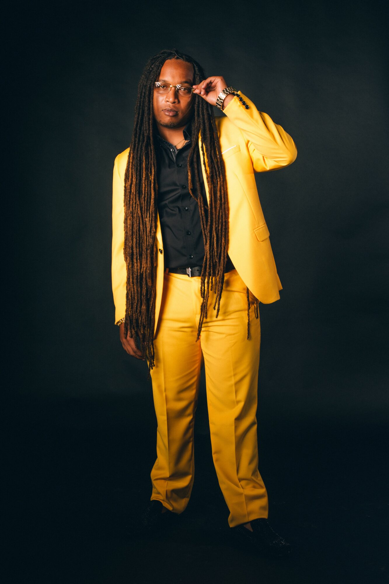 Person with long dreadlocks wearing a yellow suit and black shirt, standing against a dark background.