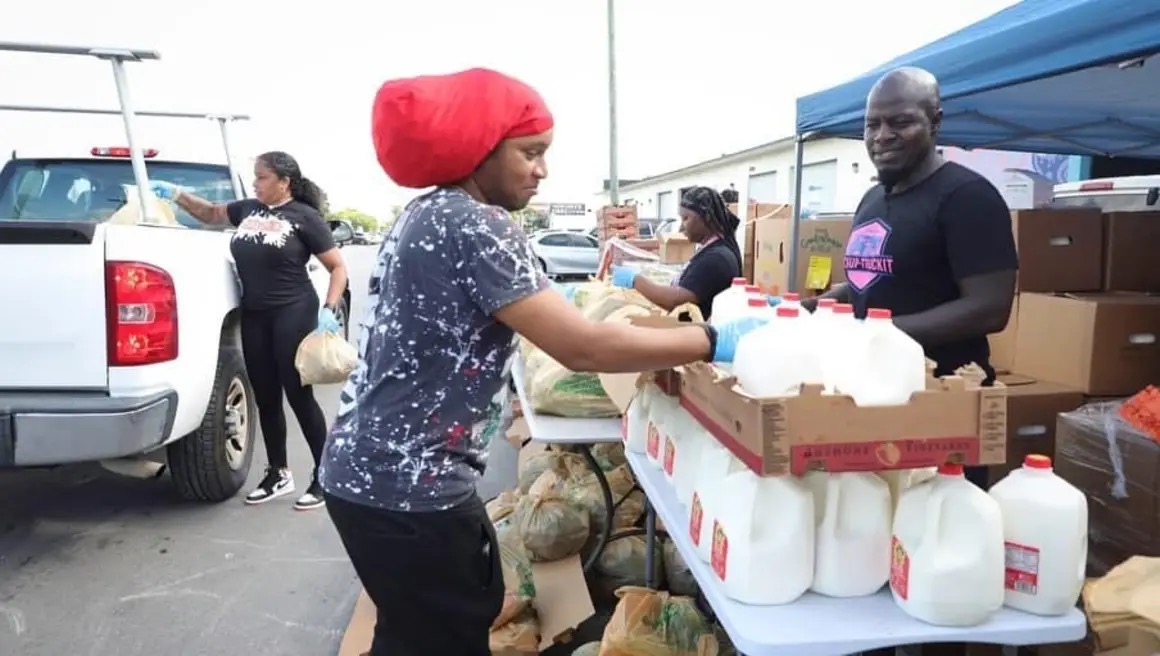 People at an outdoor market stall with milk bottles, a woman with a red headwrap, and a man behind the stall smiling.