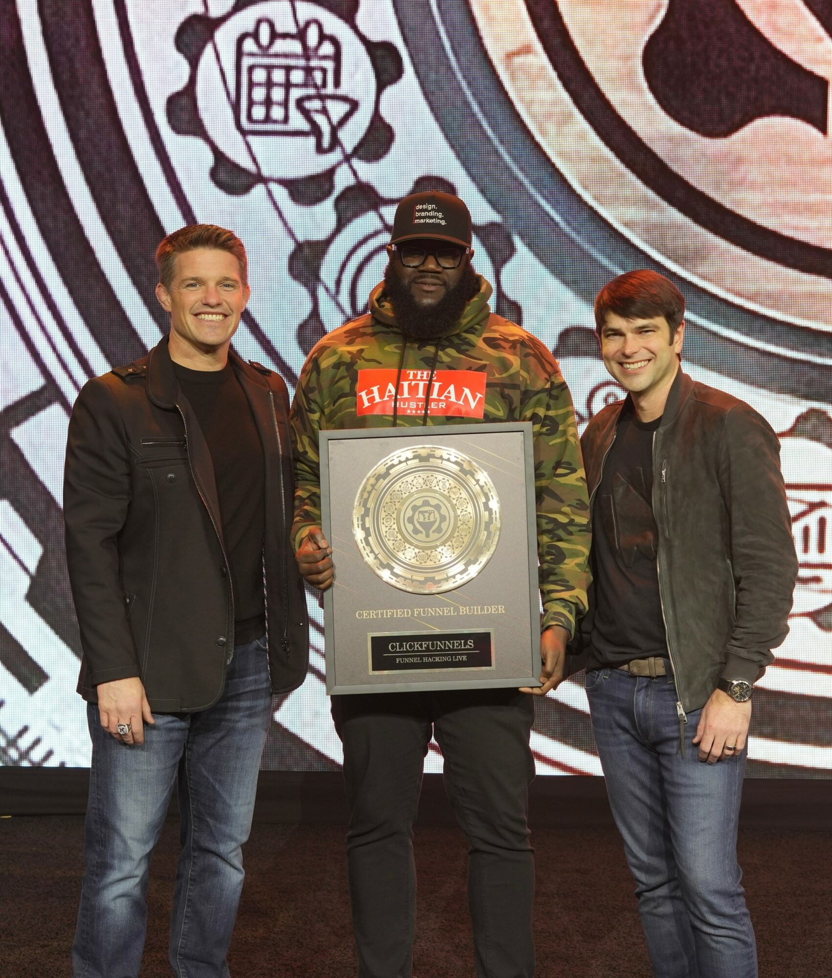 Three men standing together, one holding a framed award, smiling, in front of a patterned background.