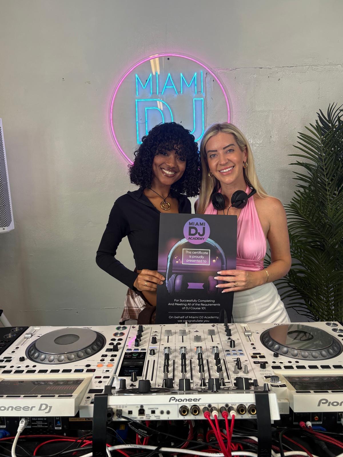 Two women stand behind DJ equipment, holding a poster, smiling, with a neon Miami sign and a plant in the background.