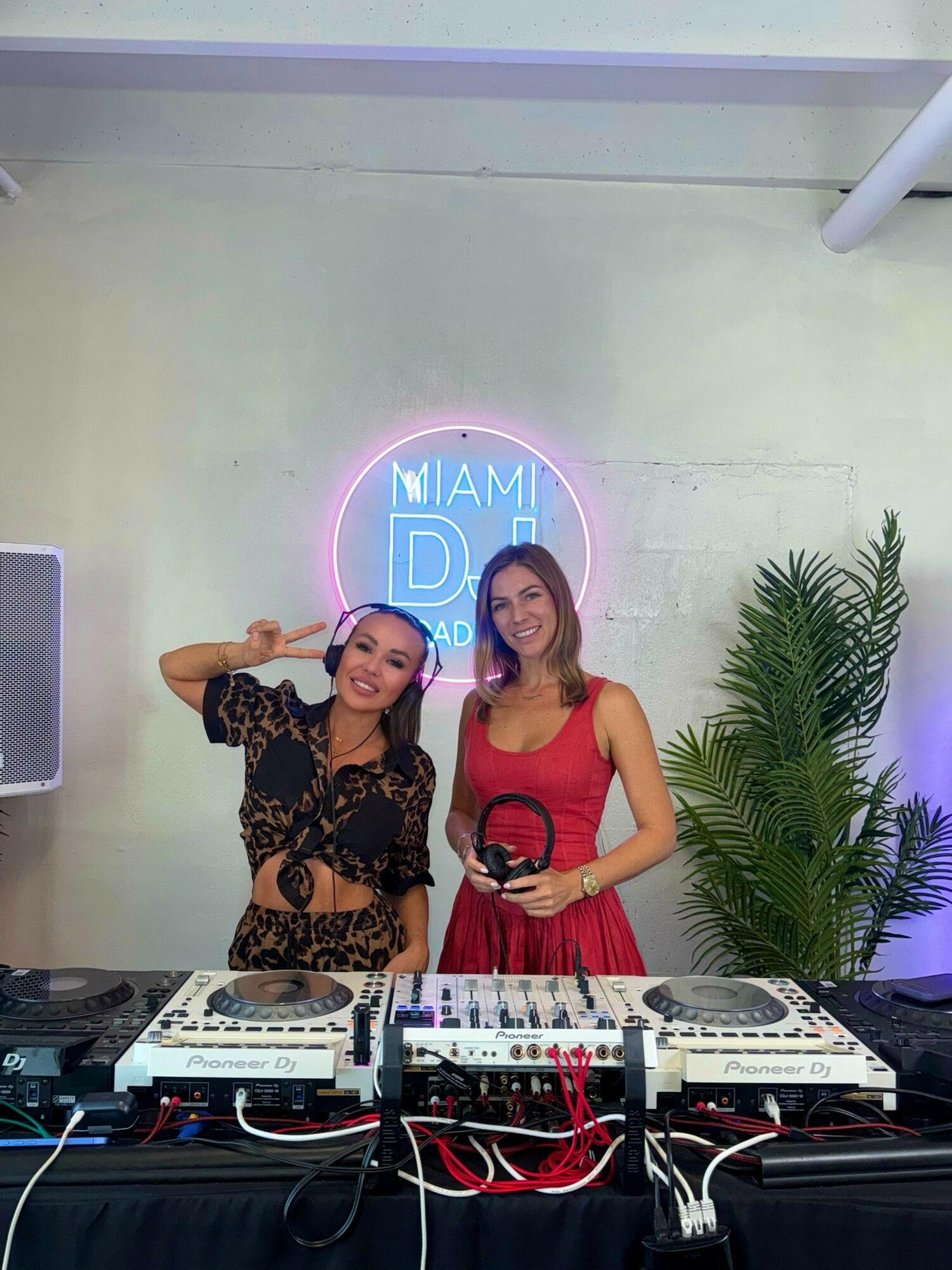 Two women stand behind DJ equipment, smiling, with a neon Miami DJ sign and a plant in the background.