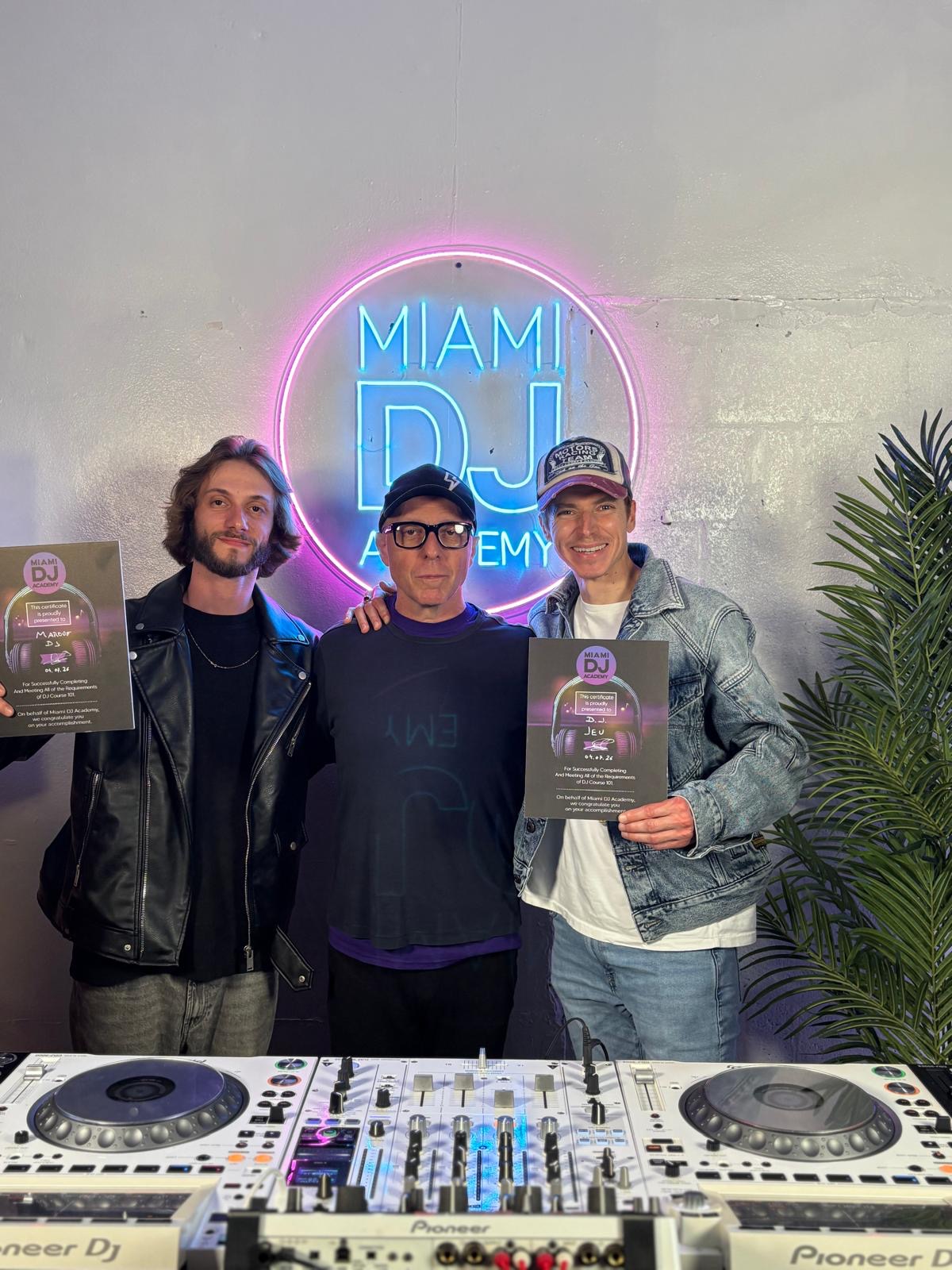 Three people stand in front of a neon sign that says 'MIAMI DJ'. Two hold posters, one wears a cap, and a DJ setup is in front.