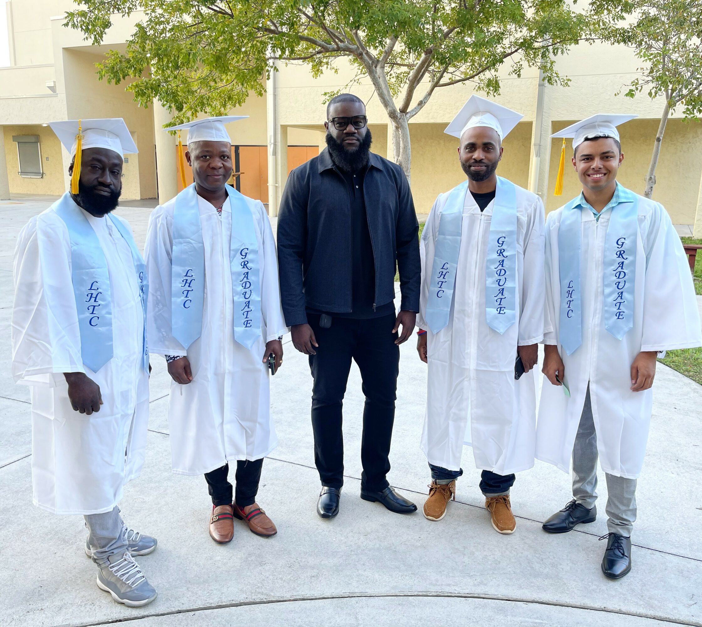 Five people standing outdoors, four in graduation gowns and caps, one in casual clothing, with trees and buildings in background.