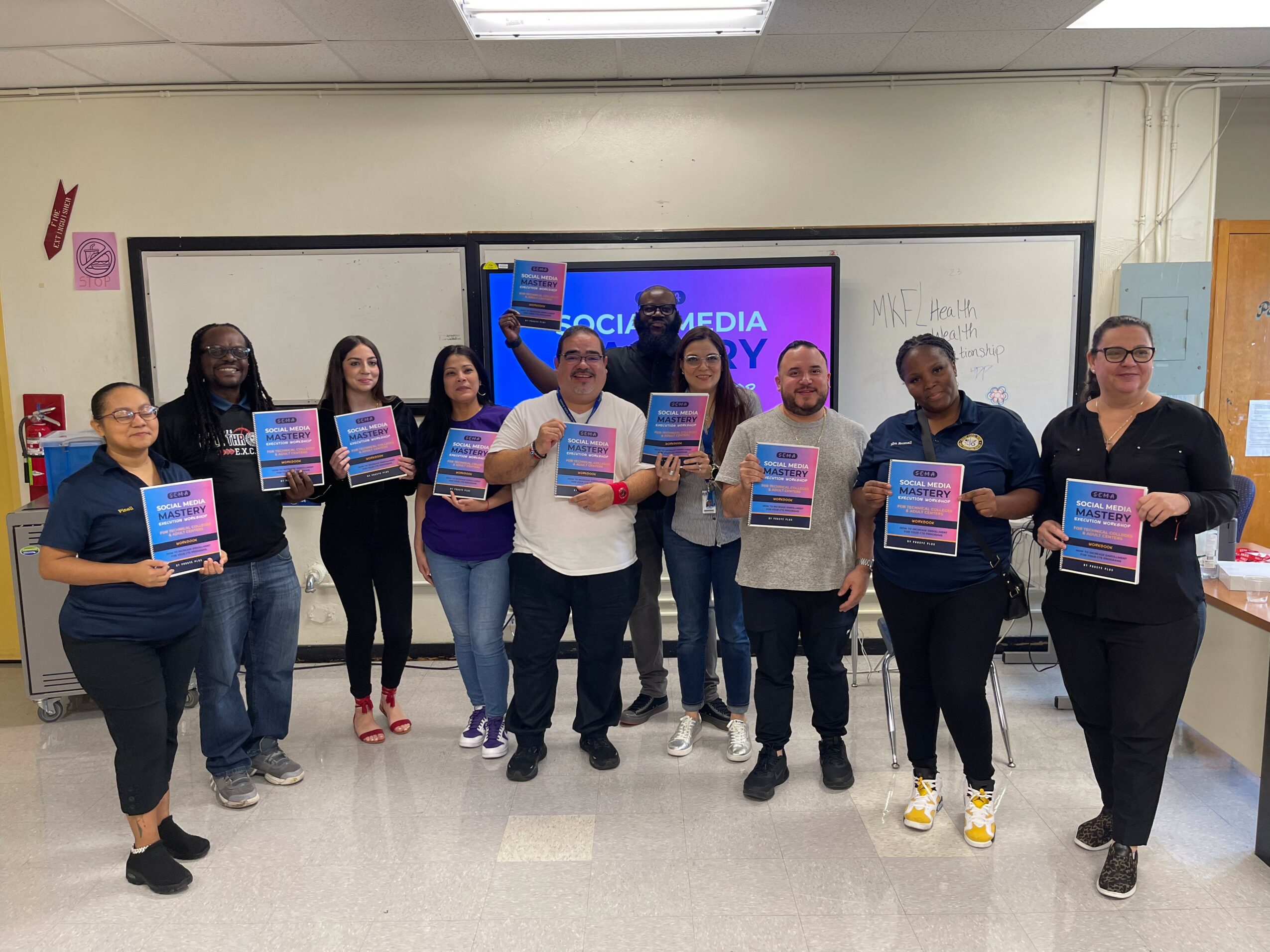 Group of ten people standing in a classroom holding certificates, with a purple and pink presentation slide on the screen behind them.