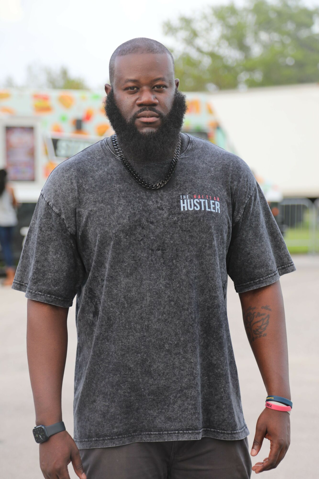 Man with a beard wearing a dark T-shirt and wristbands, standing outdoors with colorful background.