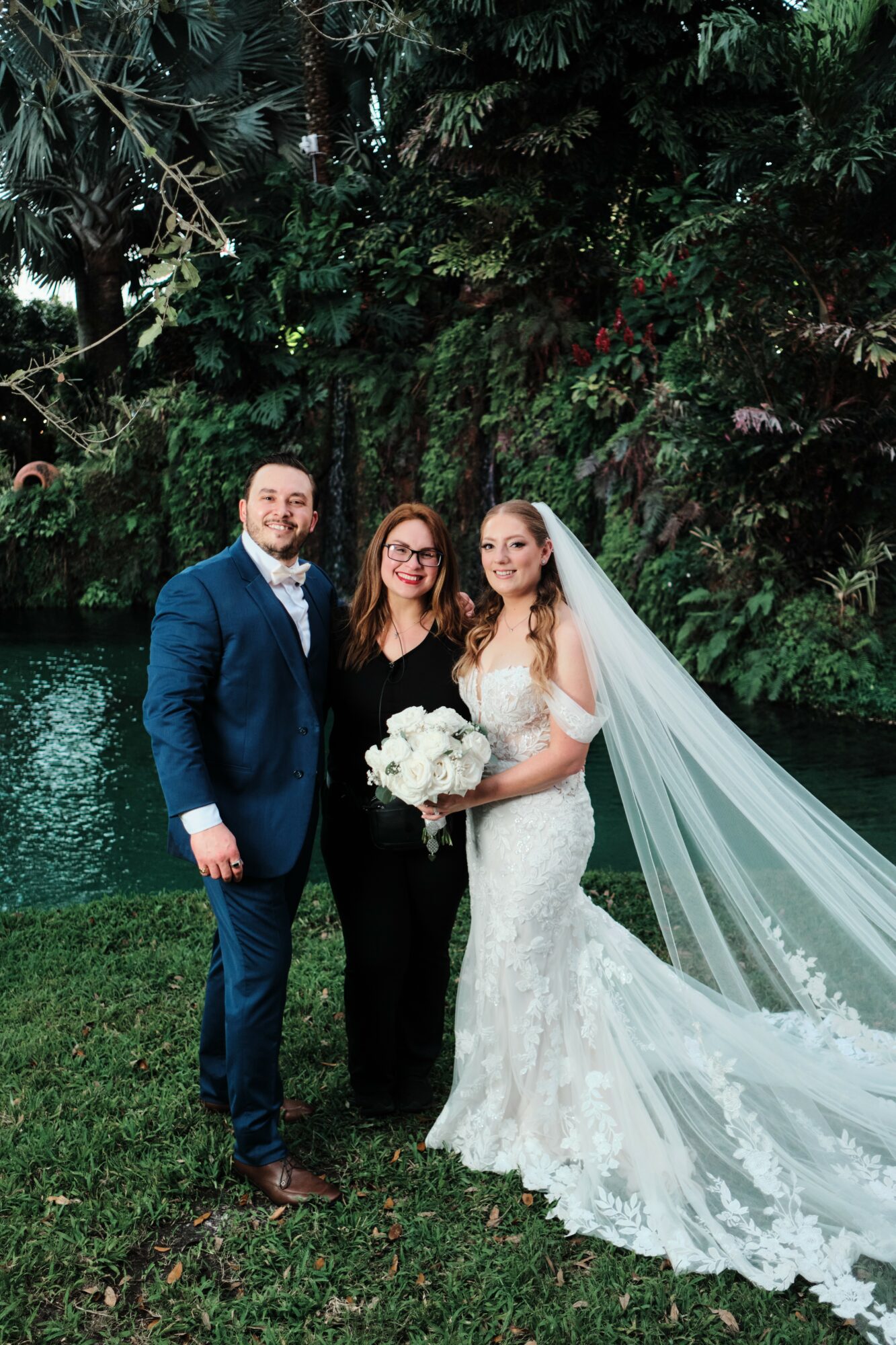 Three people standing outdoors in front of trees, one in a wedding dress holding a bouquet, two others in formal attire.