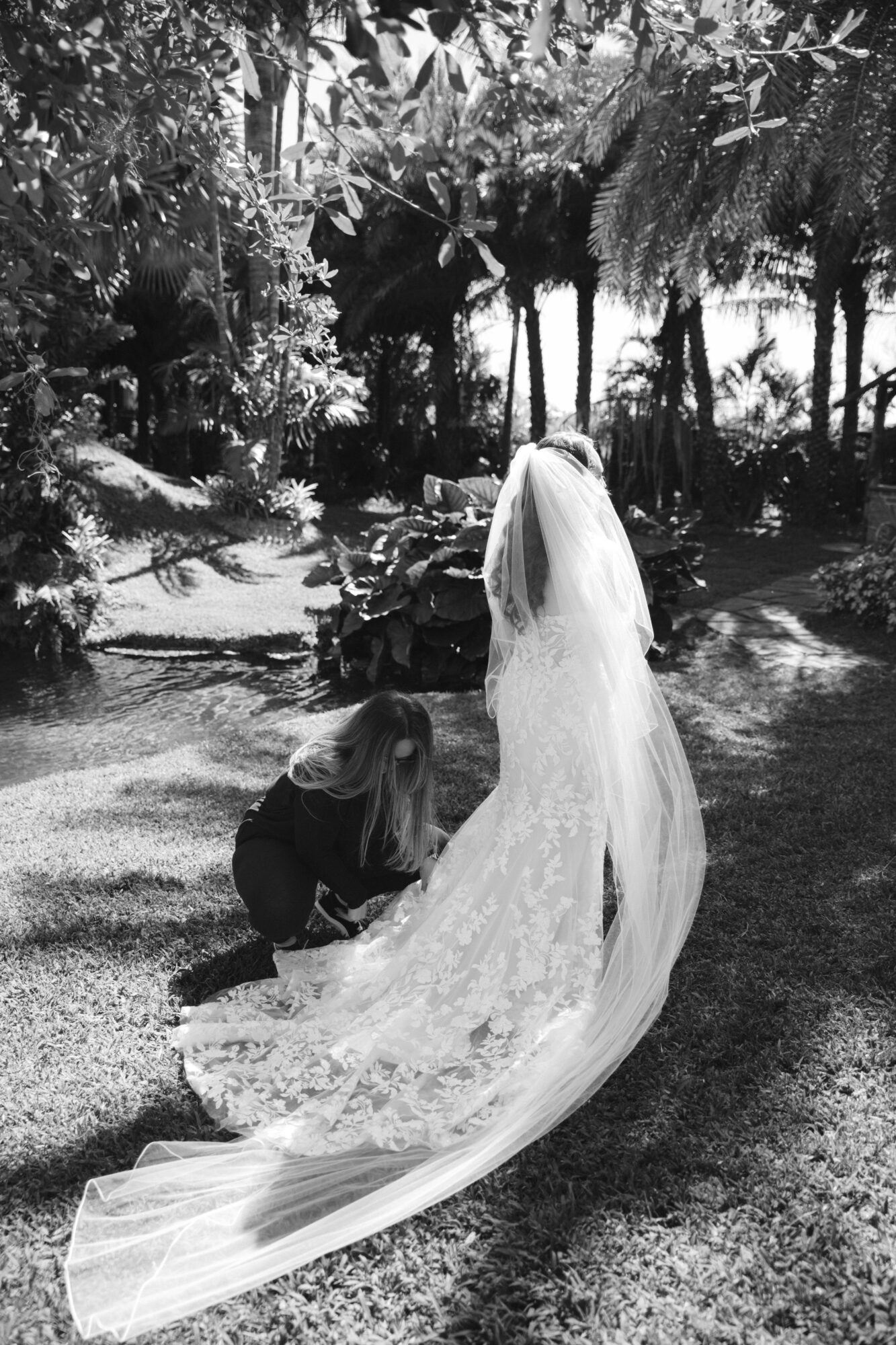 Person adjusting a long, flowing wedding dress outdoors with trees and sunlight in background.