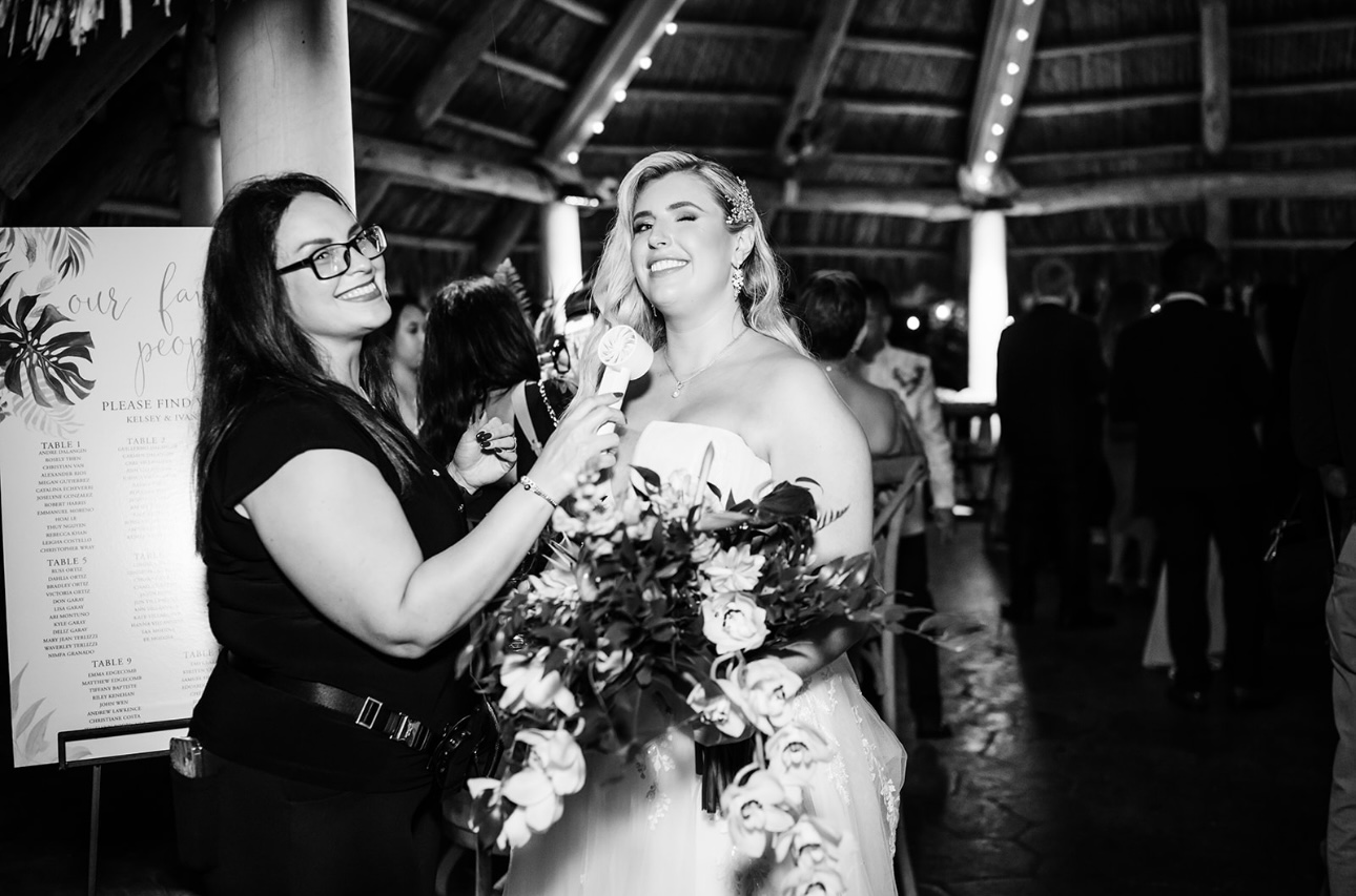 Woman in wedding dress holding flowers being interviewed by woman with microphone, group in background, black and white photo.