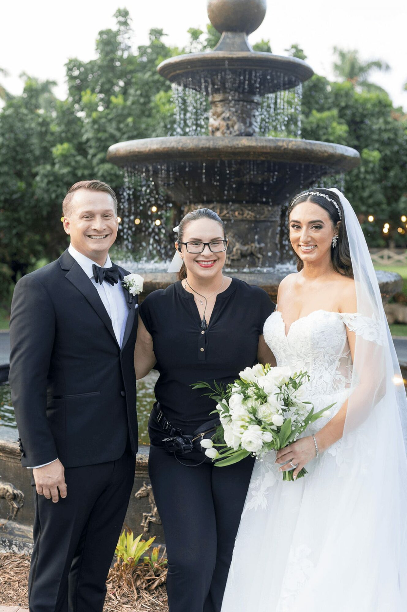 Bride in wedding dress holding bouquet, groom in tuxedo, woman in black shirt standing in front of fountain outdoors.