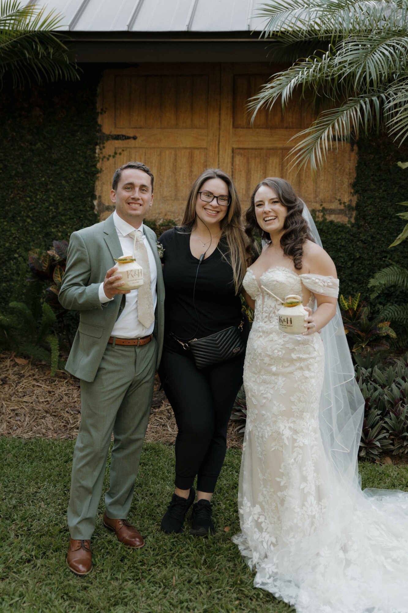 Three people standing outdoors, two women and one man, smiling and holding drinks, with greenery and a wooden door in the background.