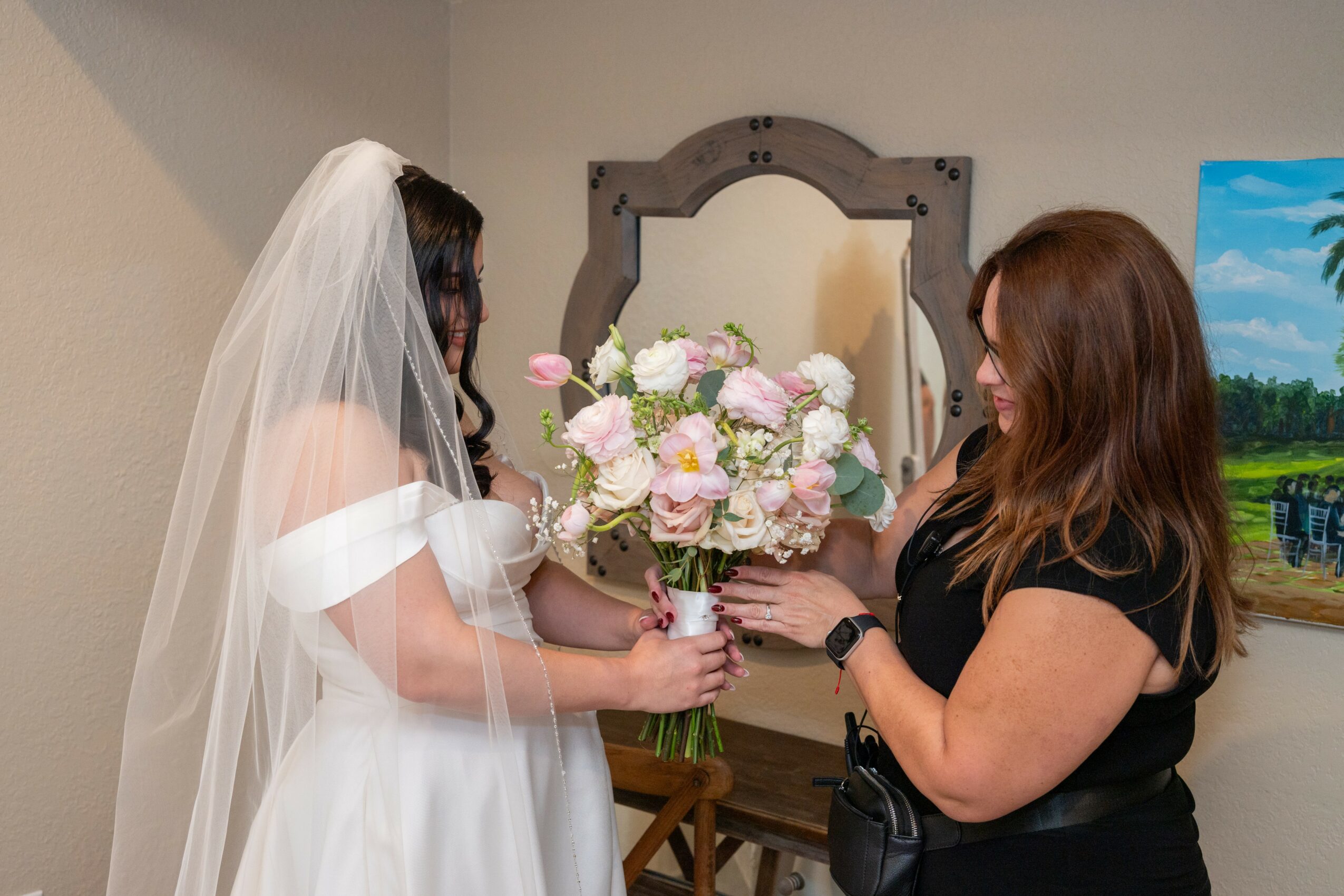 Two women exchanging a bouquet of pink and white flowers indoors, one wearing a wedding dress and veil, the other in black, with a mirror and painting in the background.