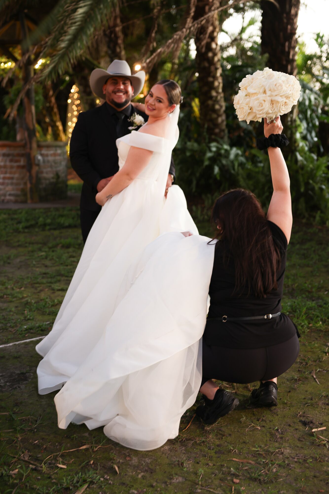 Bride in white dress holding bouquet, man in cowboy hat, woman kneeling, outdoors with trees and sunlight, wedding scene