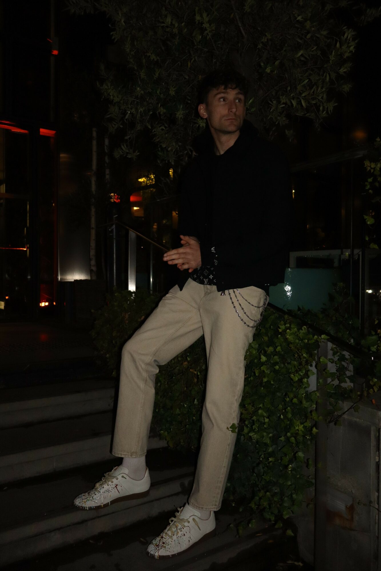 Person standing outdoors at night, wearing a black jacket, beige pants, and white shoes, holding an object in hand.