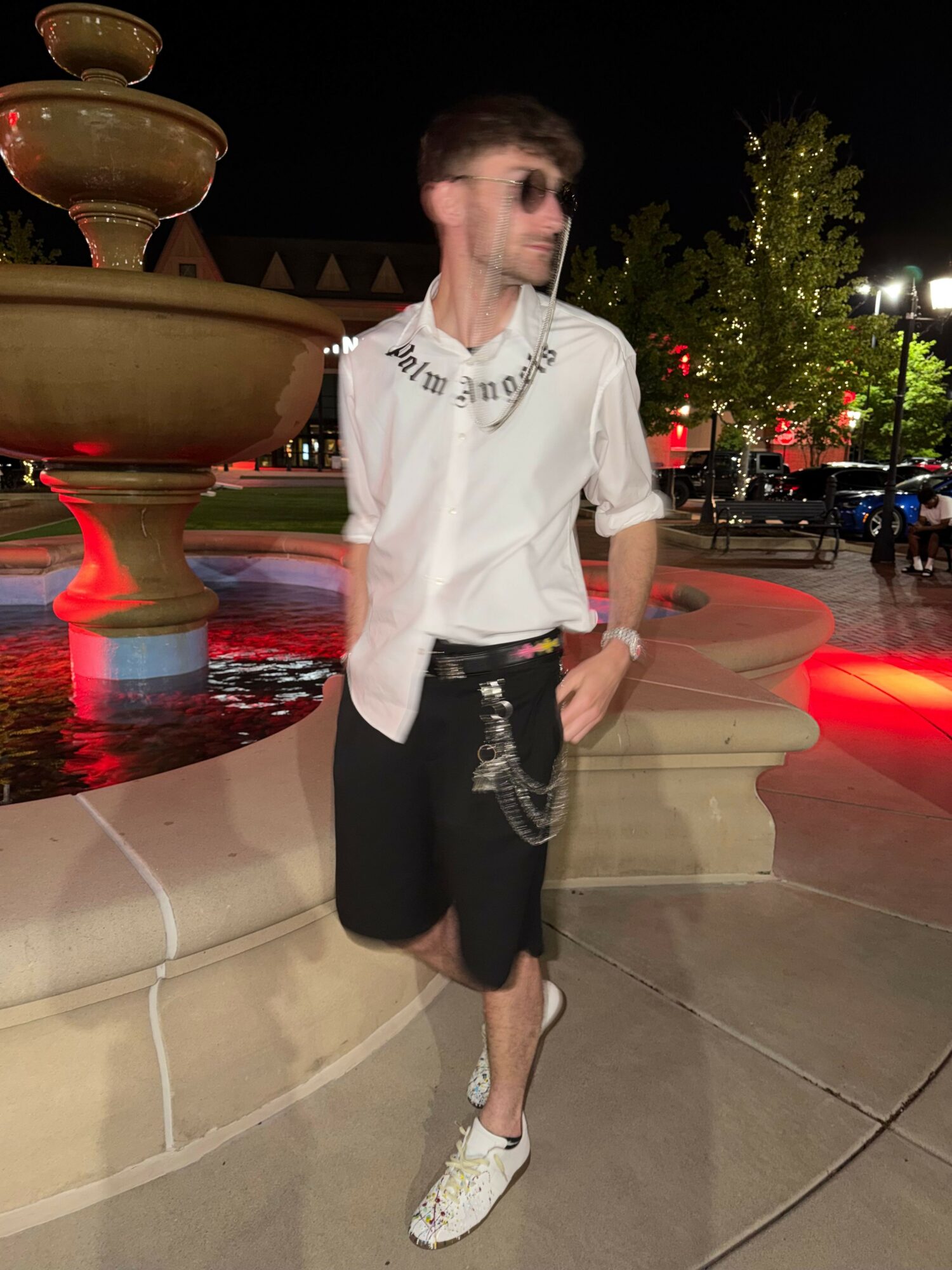 Person standing outdoors at night, wearing a white shirt, black skirt, and white shoes, with trees and lights in the background.