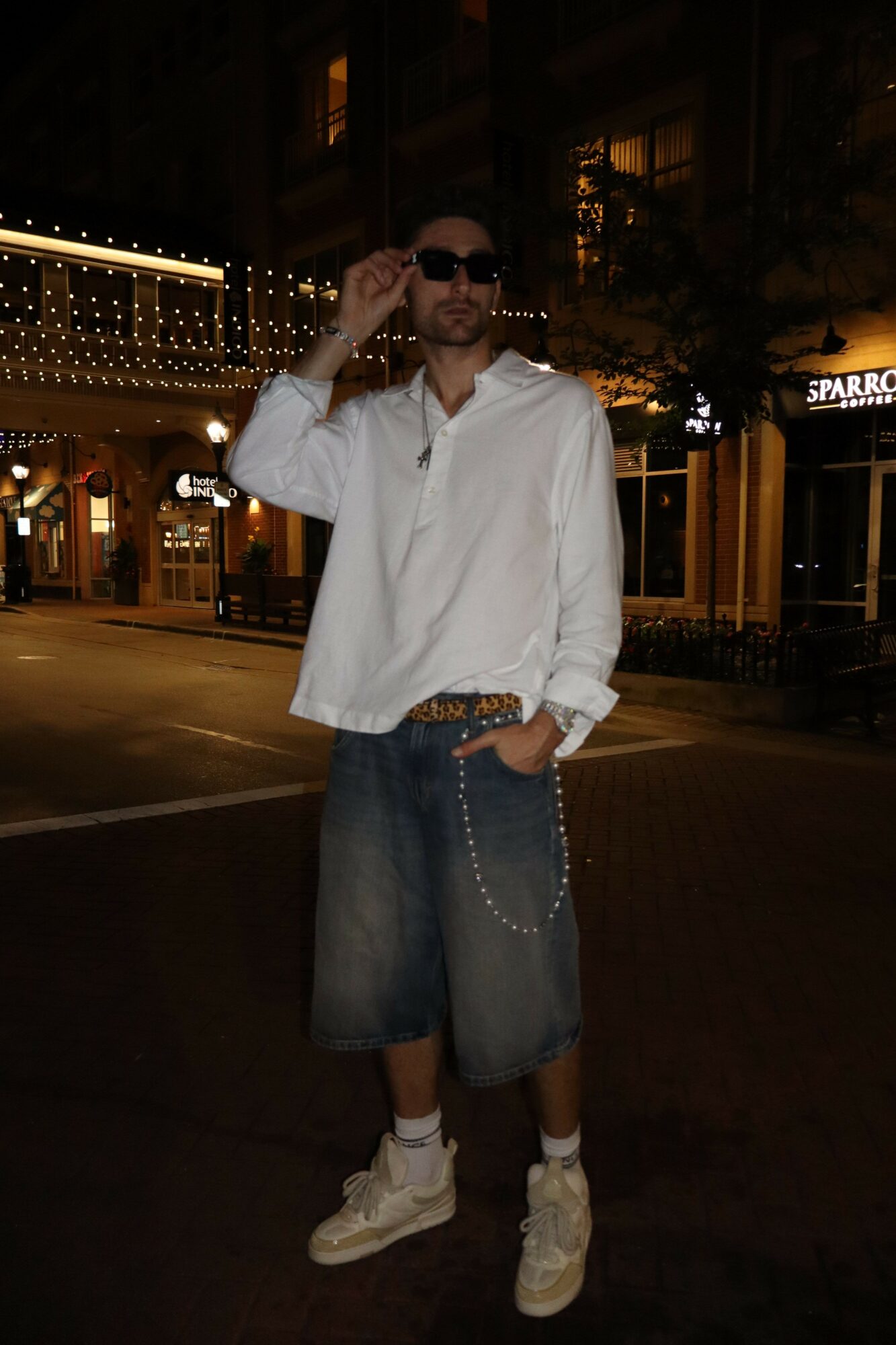 Person standing outdoors at night, wearing sunglasses, white shirt, jeans, and sneakers, holding a phone to their ear.