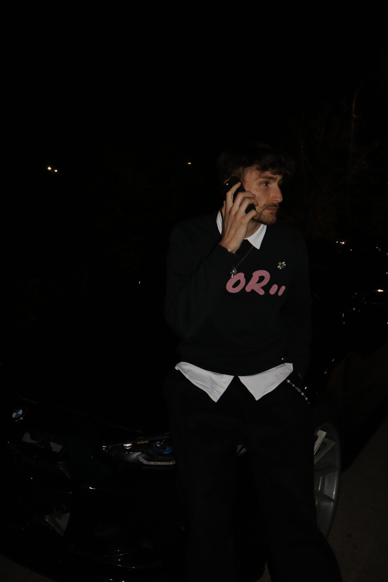Person standing outdoors at night, talking on a phone, with dark background and faint lights, wearing dark clothing.