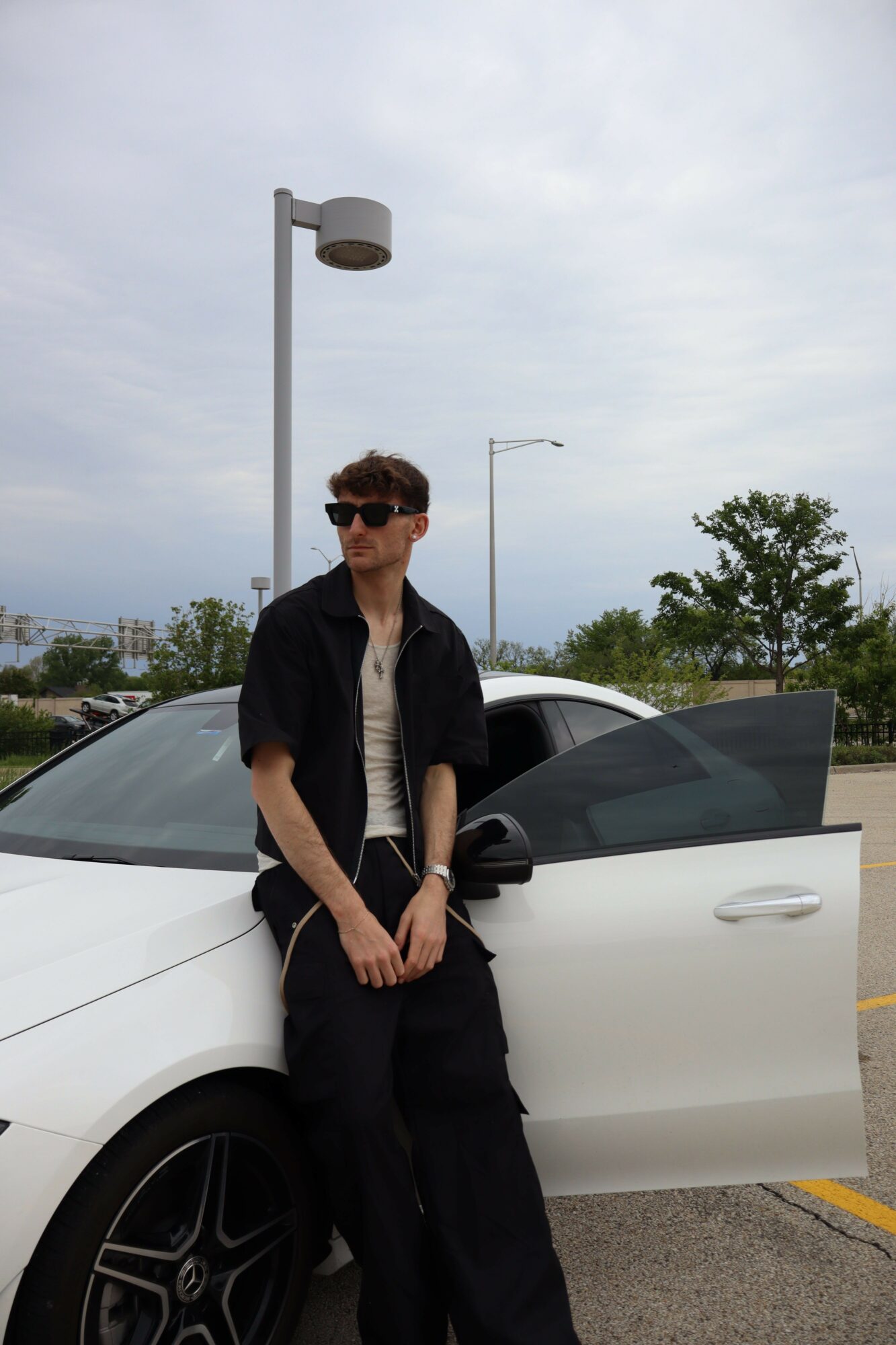 Person with short hair and sunglasses leaning against a white car with an open door, outdoors on a cloudy day.