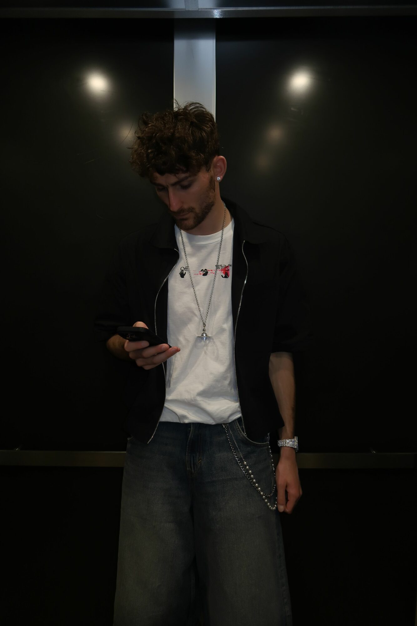 Person standing in a dark room, looking at a phone, wearing a white shirt and dark jacket, with a watch on wrist.