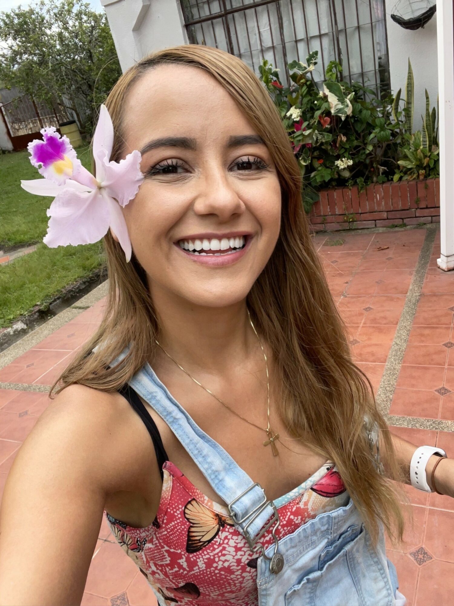 Young woman smiling outdoors with a flower in her hair, wearing a floral top and denim overalls, in a garden setting.