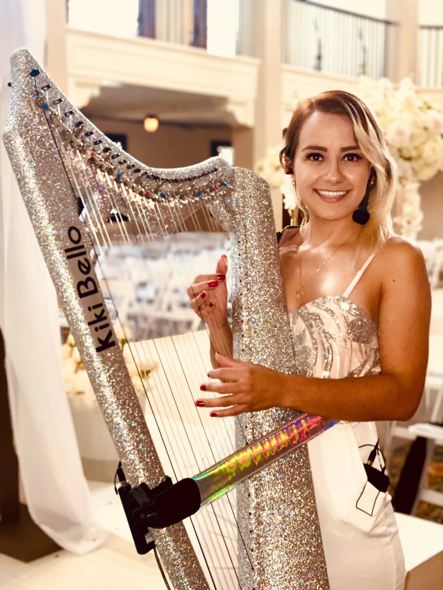 Young woman with a harp, smiling, in an elegant indoor setting with flowers and a balcony, wearing a dress with spaghetti straps.
