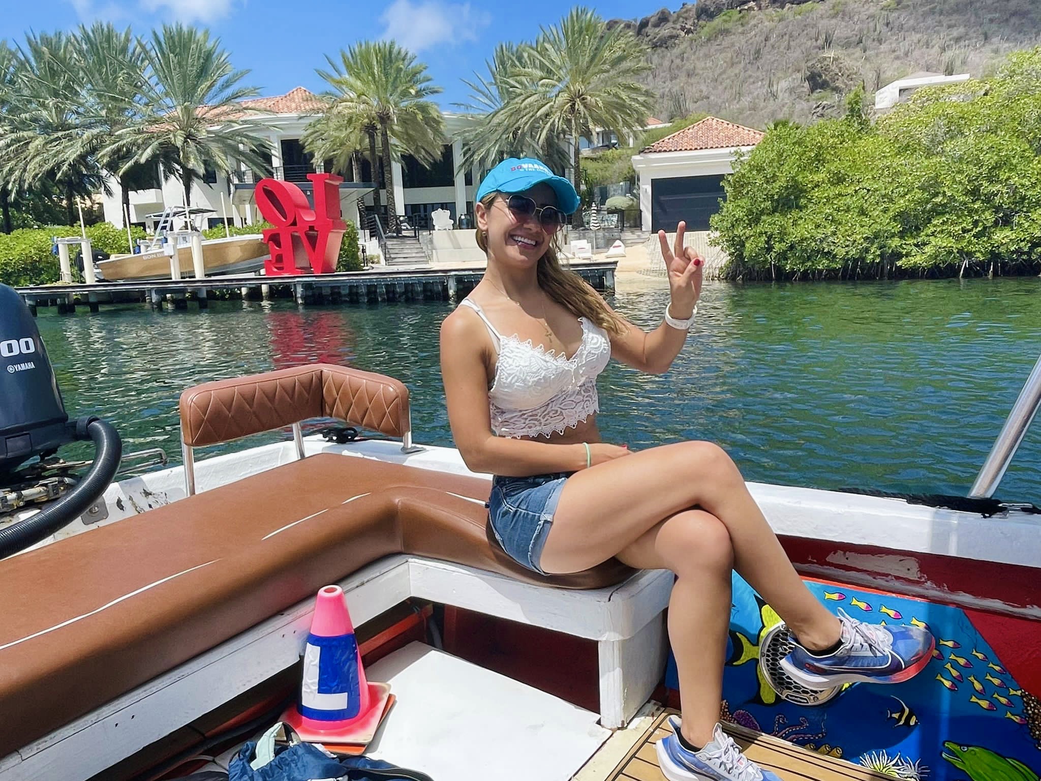 Woman sitting on boat with water, trees, and buildings in background, waving and smiling, wearing a cap, tank top, and shorts.
