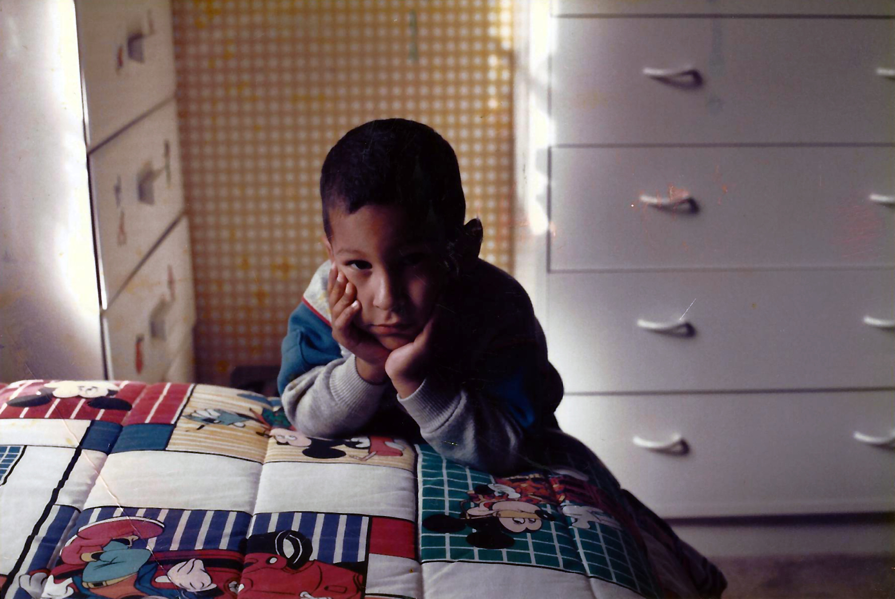 Child lying on bed with head resting on hands, smiling, in a room with dresser and patterned wall behind.