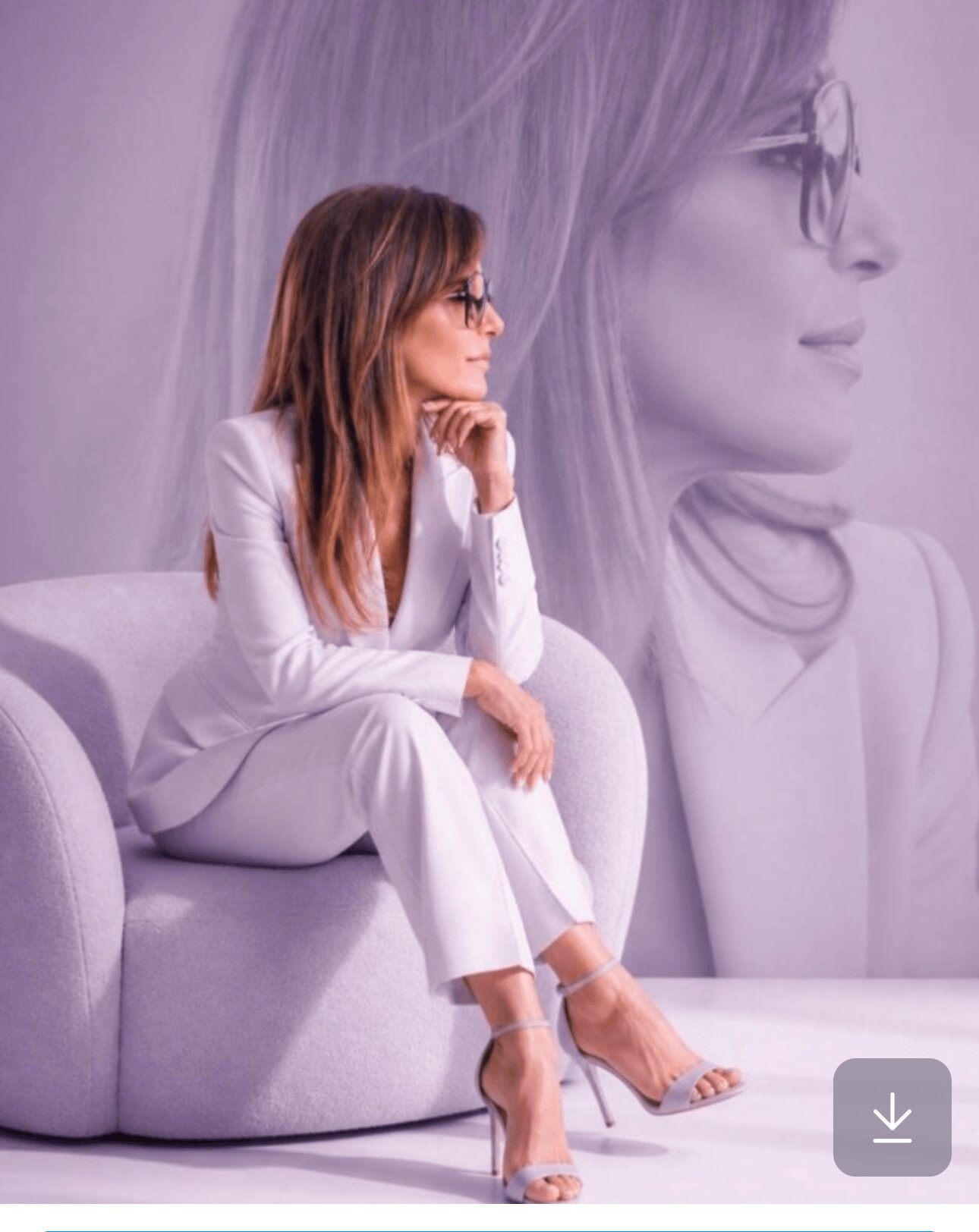 Woman with glasses sitting on a white sofa, large black-and-white portrait of her in the background, wearing white outfit.