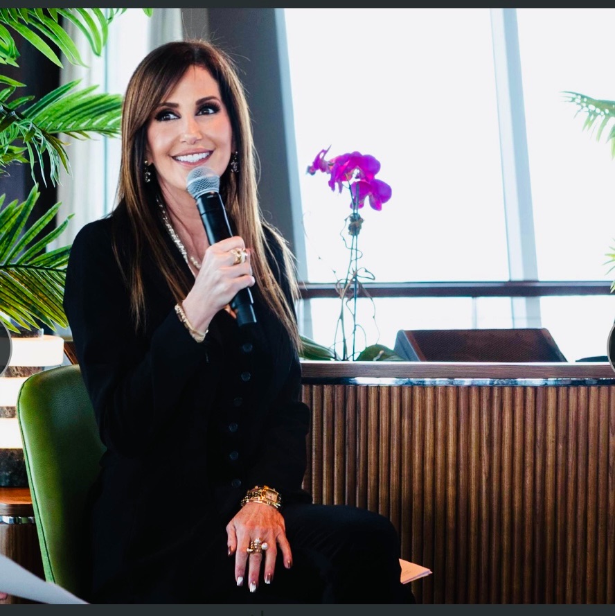 Woman with long hair holding a microphone, sitting indoors with plants and a window in the background.