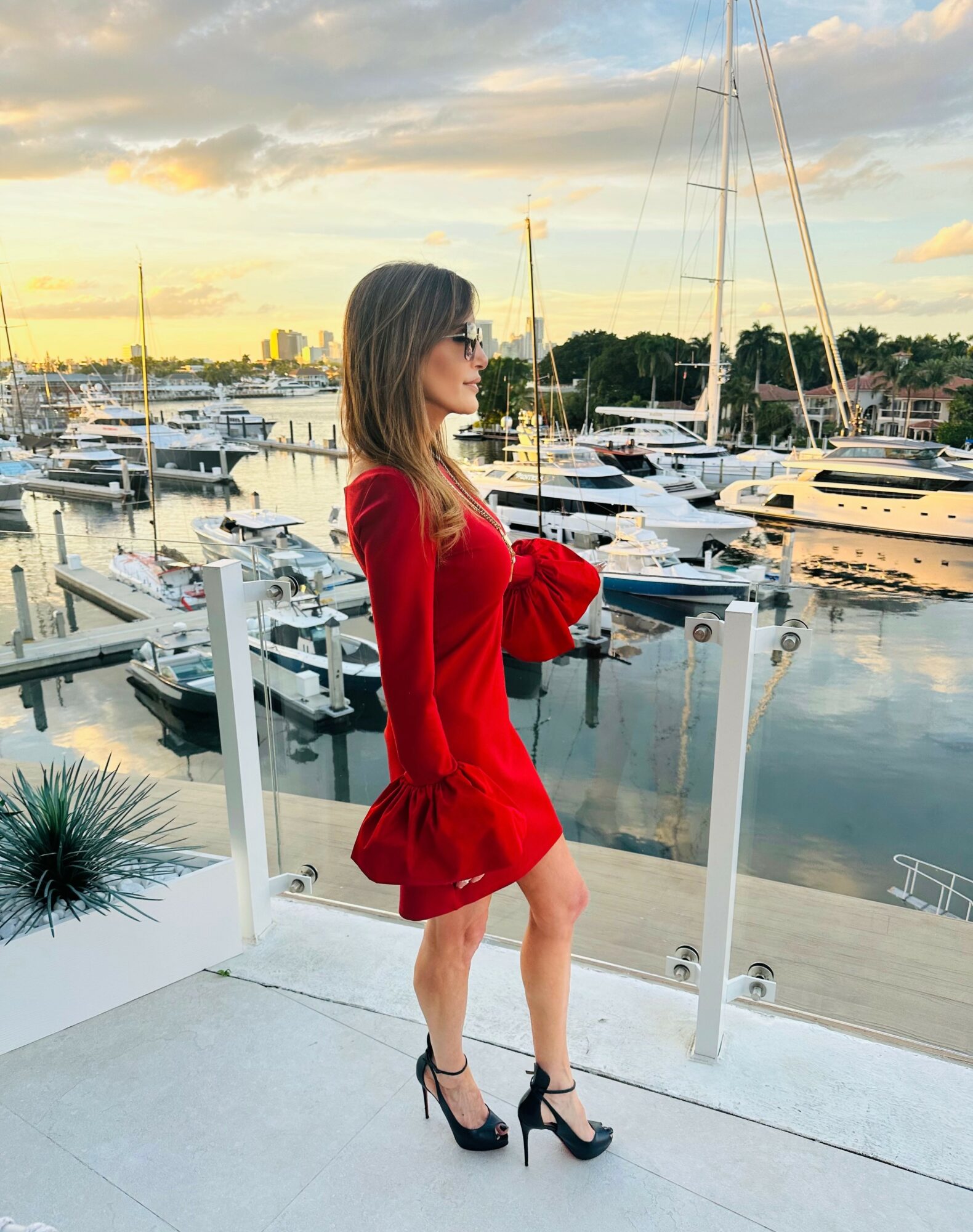 Woman in red dress and high heels standing on a balcony overlooking a marina with boats and yachts at sunset.
