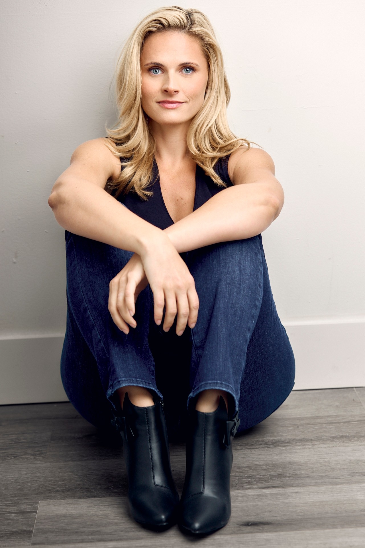 Woman with blonde hair sitting against a wall, wearing a sleeveless top, jeans, and black boots, smiling softly.