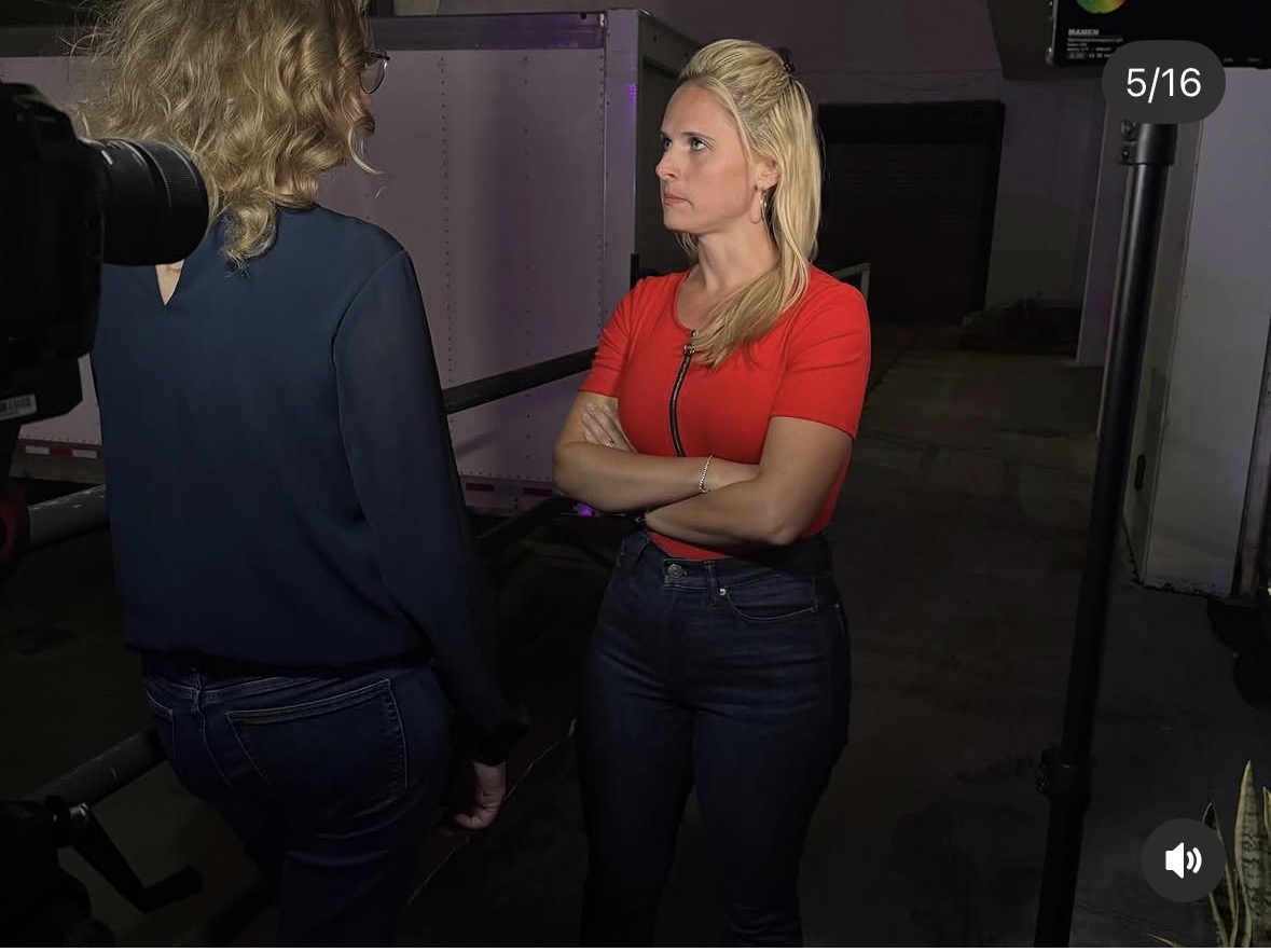 Two women standing and talking indoors, one with arms crossed, in a dimly lit room.