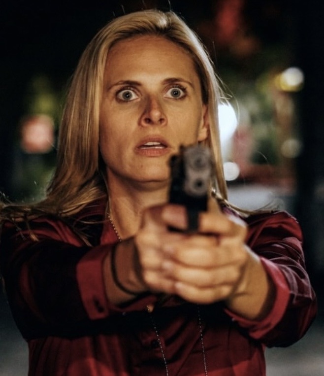 Woman with blonde hair holding a gun with both hands, aiming forward, with a surprised expression, outdoors at night.