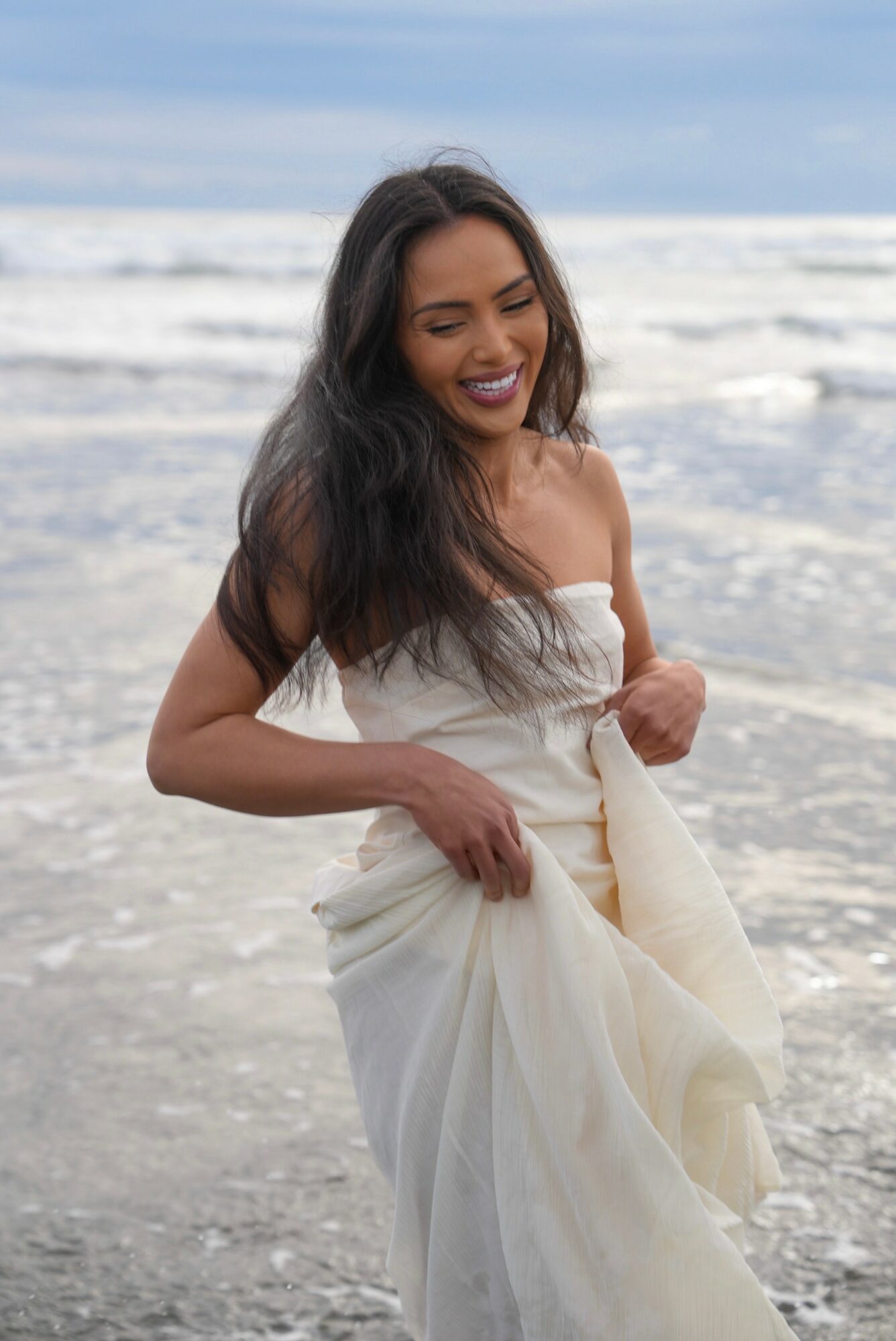 Smiling woman in a white dress standing in shallow ocean water with waves behind her.