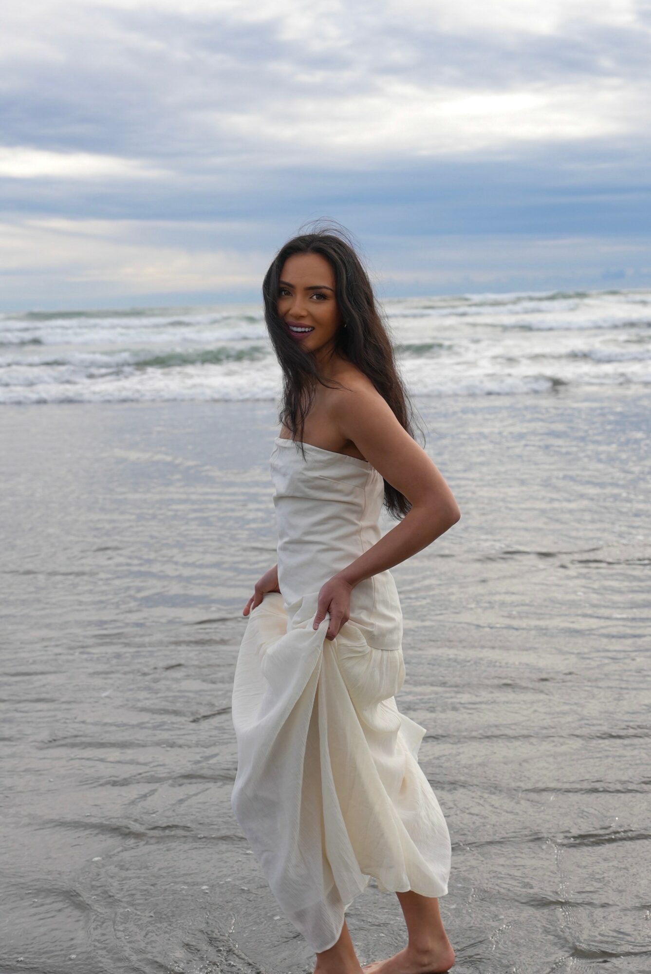Woman in white dress standing on beach with ocean and cloudy sky background, smiling, holding her dress, facing camera.