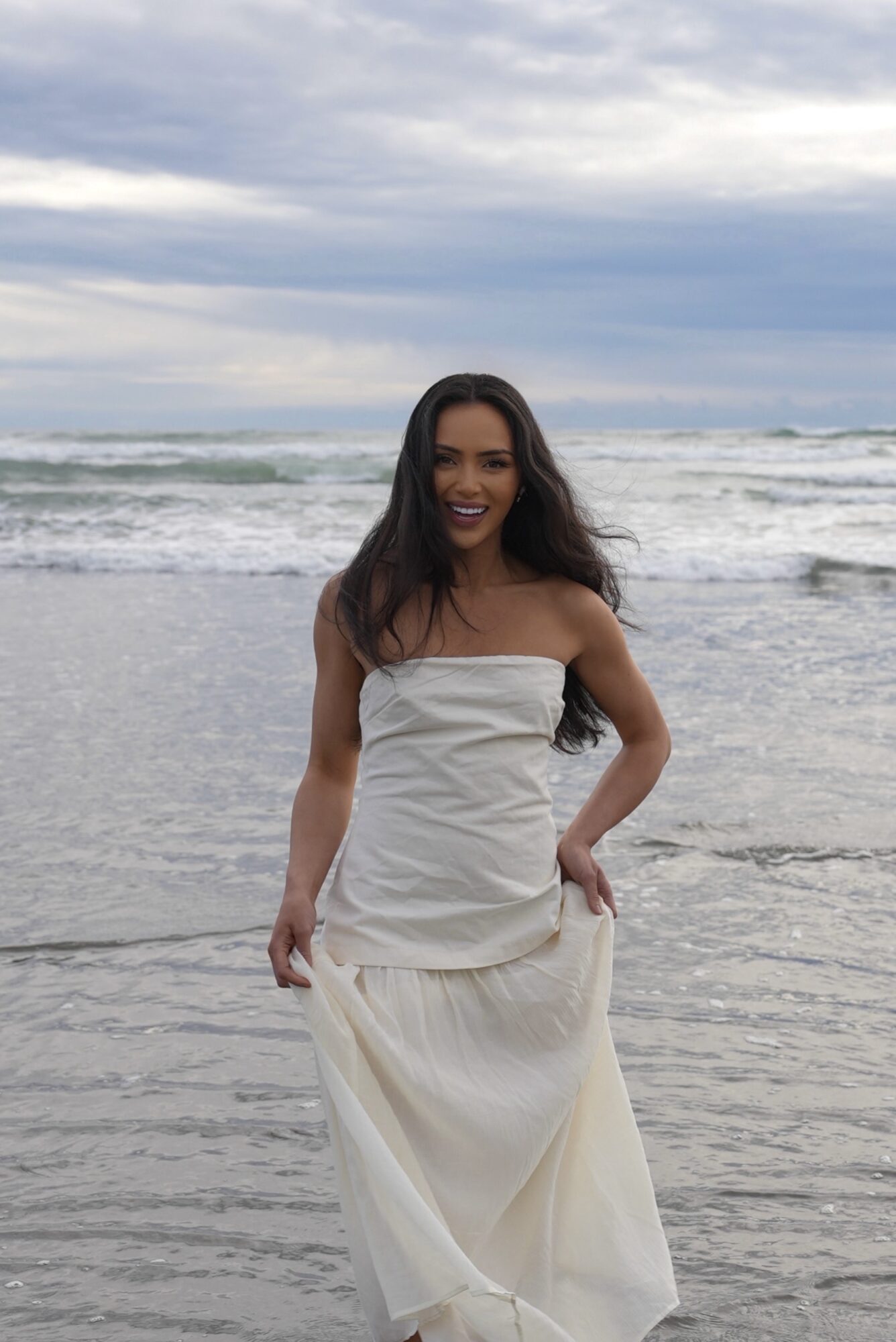 Woman in white dress standing on beach with ocean and cloudy sky in background.