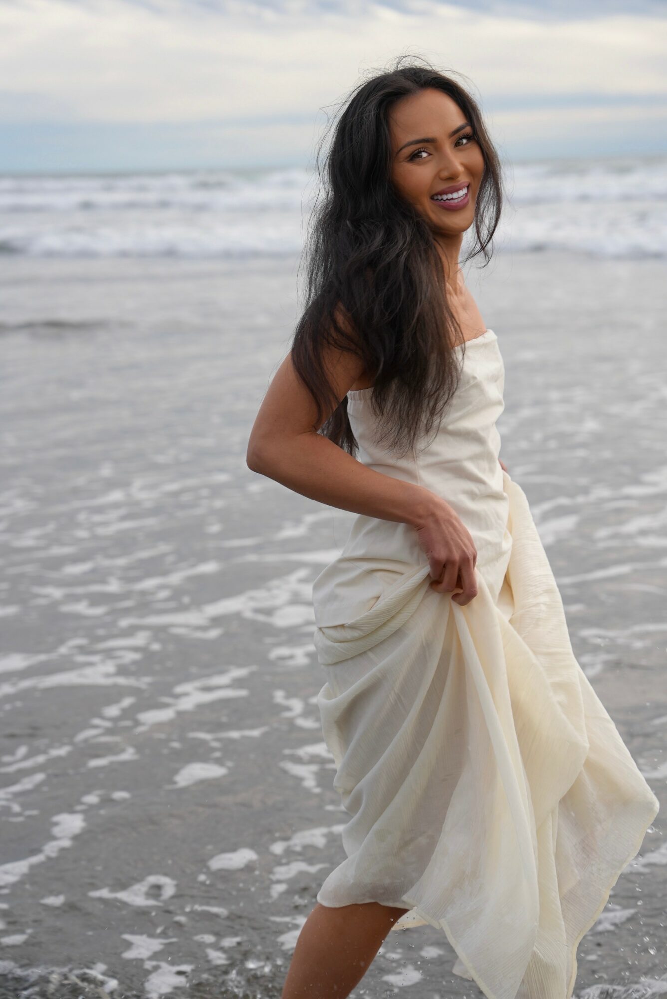 Woman in a white dress standing on the beach with ocean waves behind her, smiling and looking at the camera.