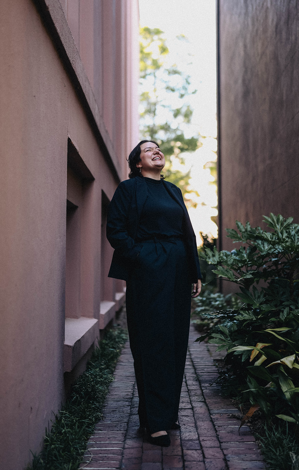 Person standing on a brick pathway between tall buildings, smiling with eyes closed, surrounded by greenery.