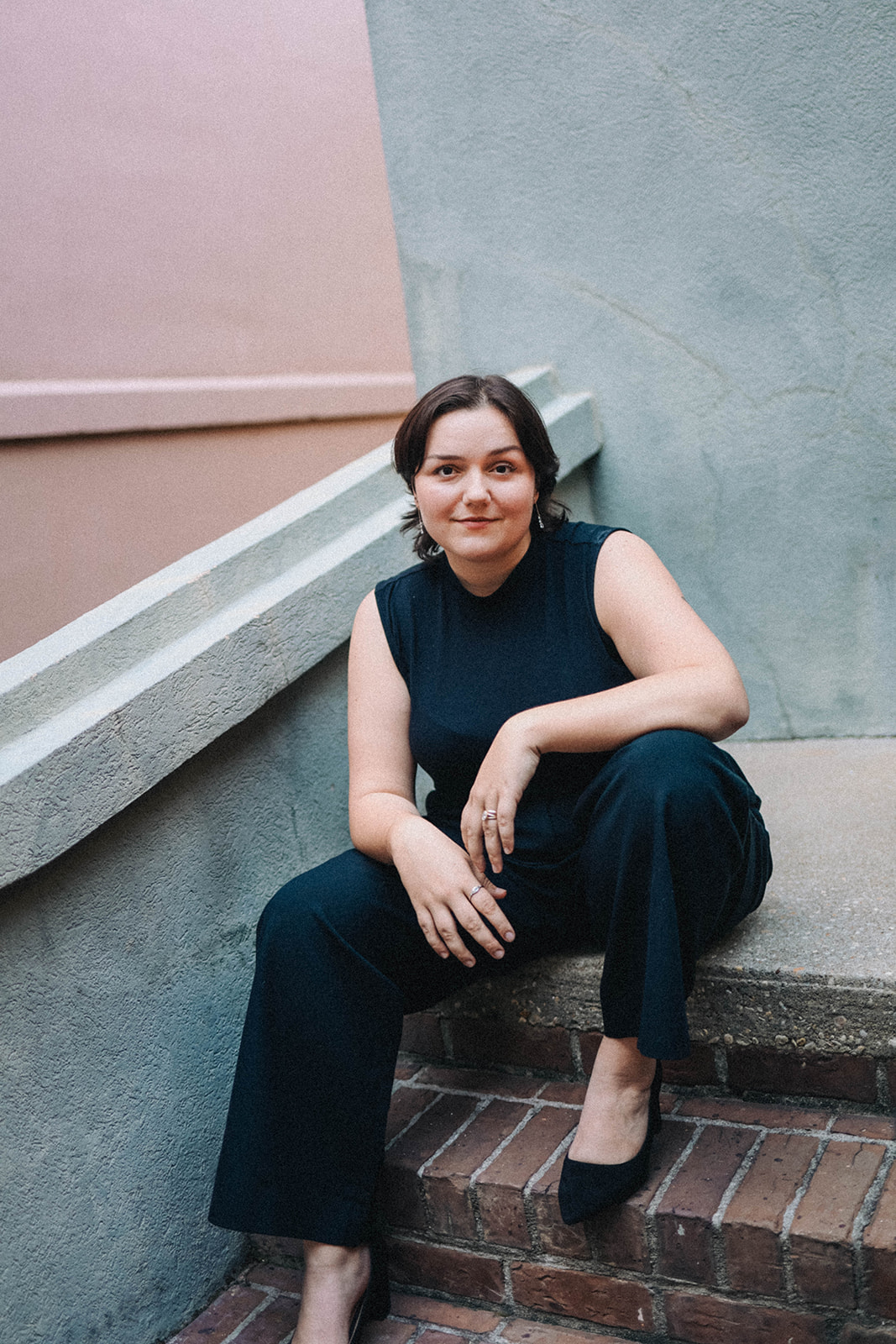 Woman with short dark hair sitting on stairs, wearing a sleeveless black top and black pants, smiling.