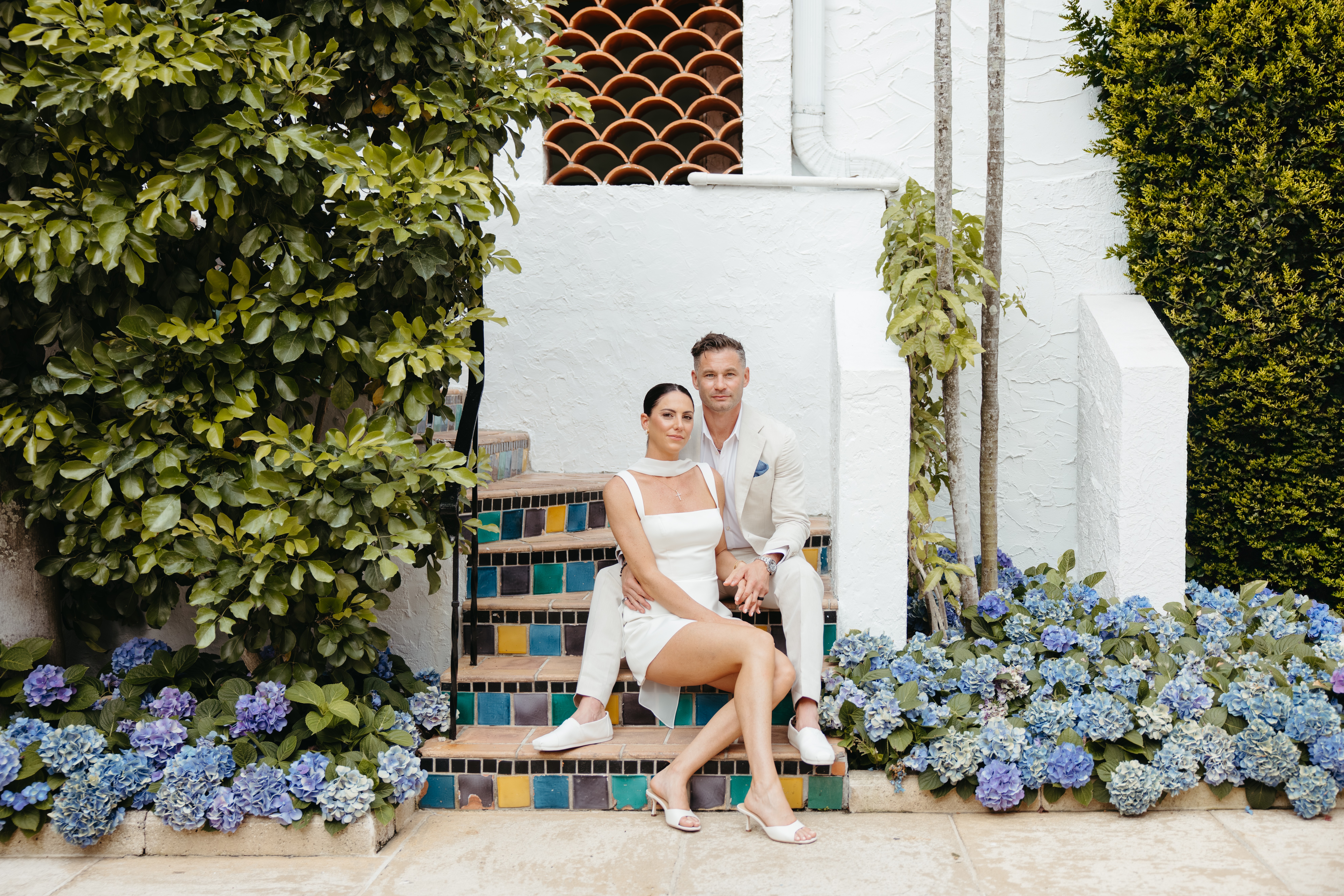 Two people sitting on colorful tiled stairs surrounded by lush greenery and blue hydrangea flowers.