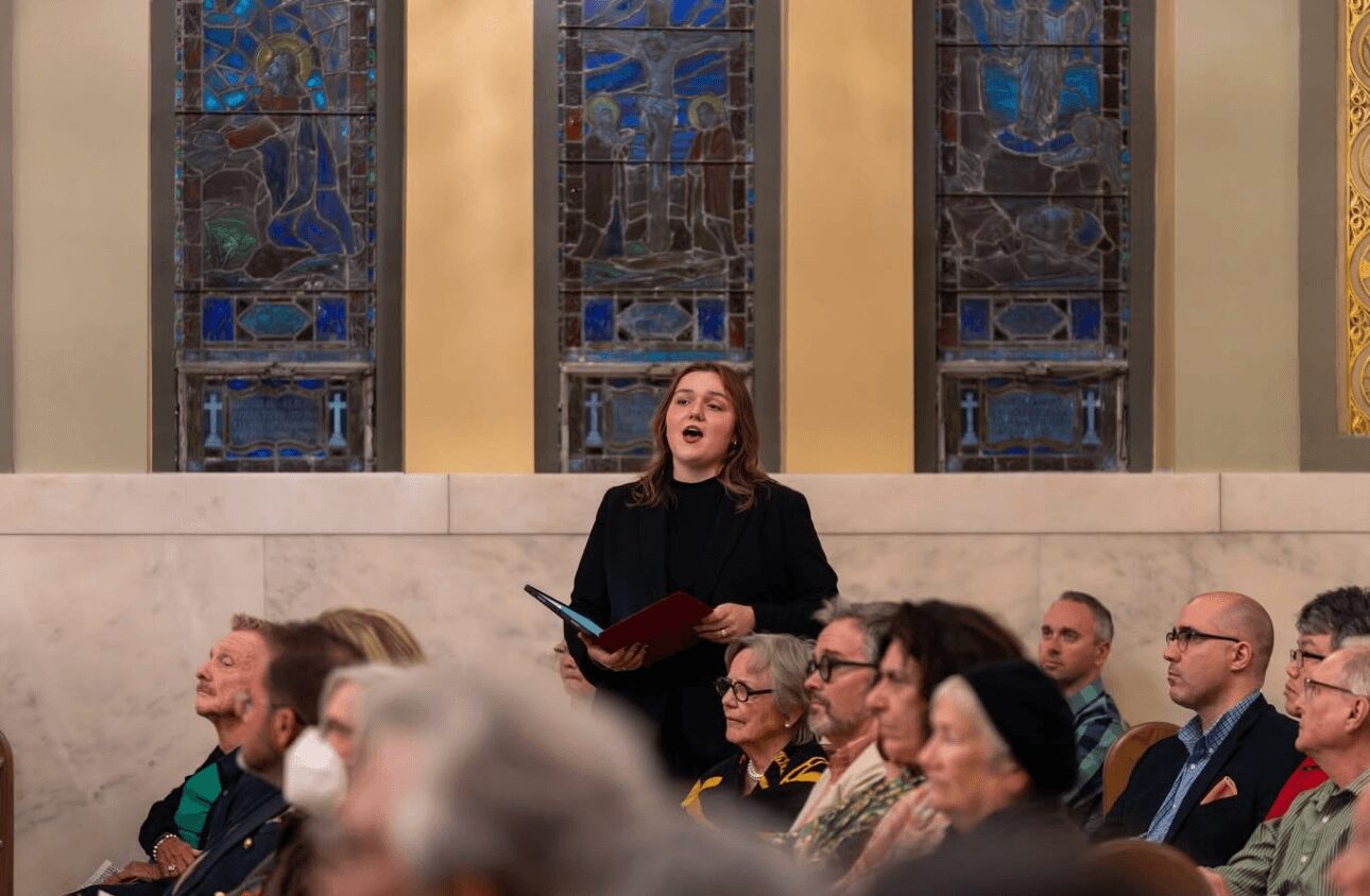 Woman singing or speaking in a church or cathedral with stained glass windows behind her, surrounded by seated audience.