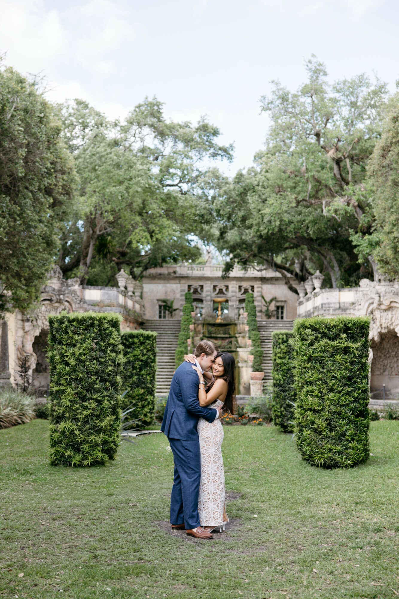 A couple stands close together in a garden with trimmed bushes and large trees, embracing and kissing.