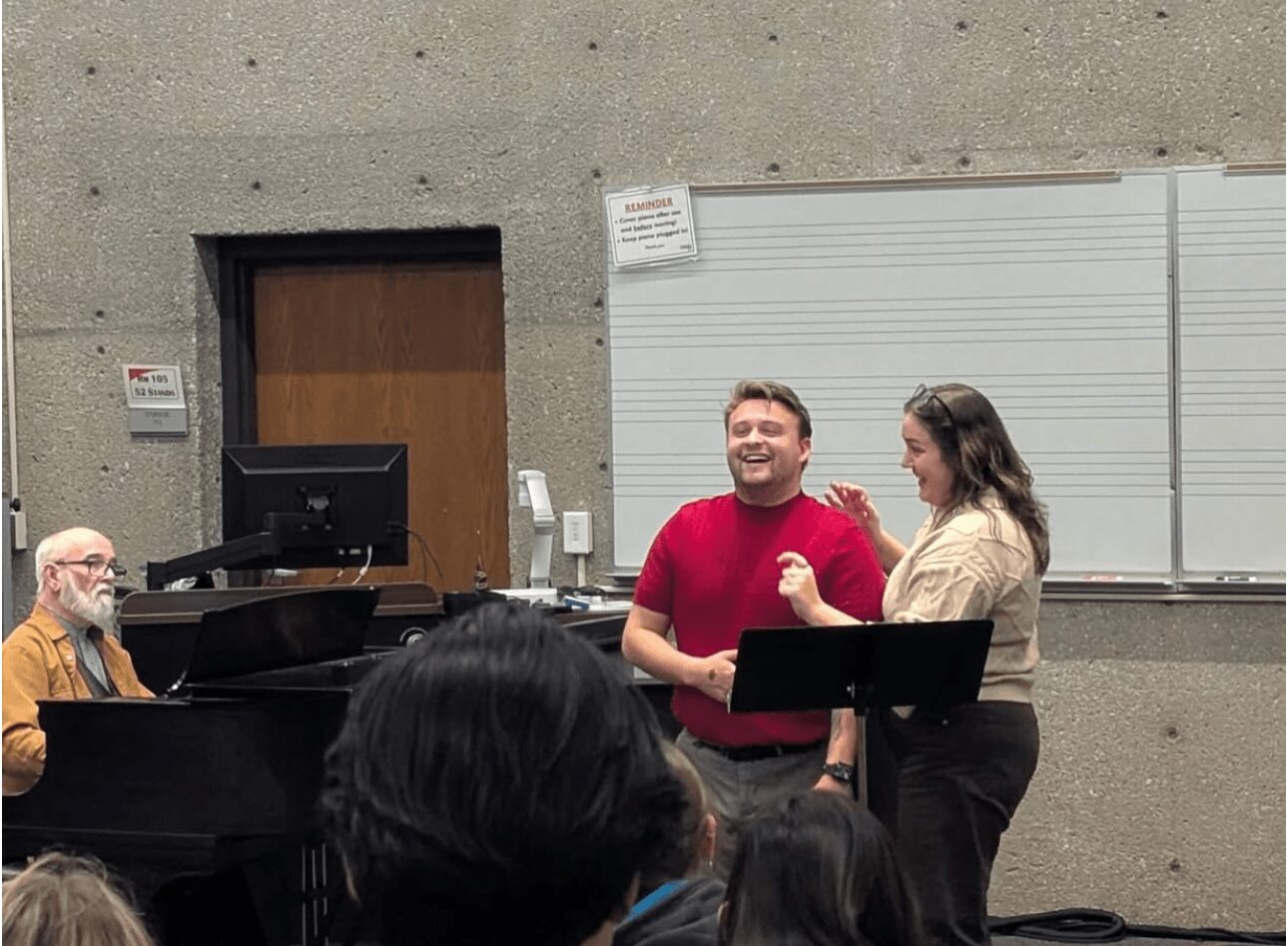 Two people stand and talk in front of a classroom whiteboard, with a pianist playing nearby and audience in foreground.
