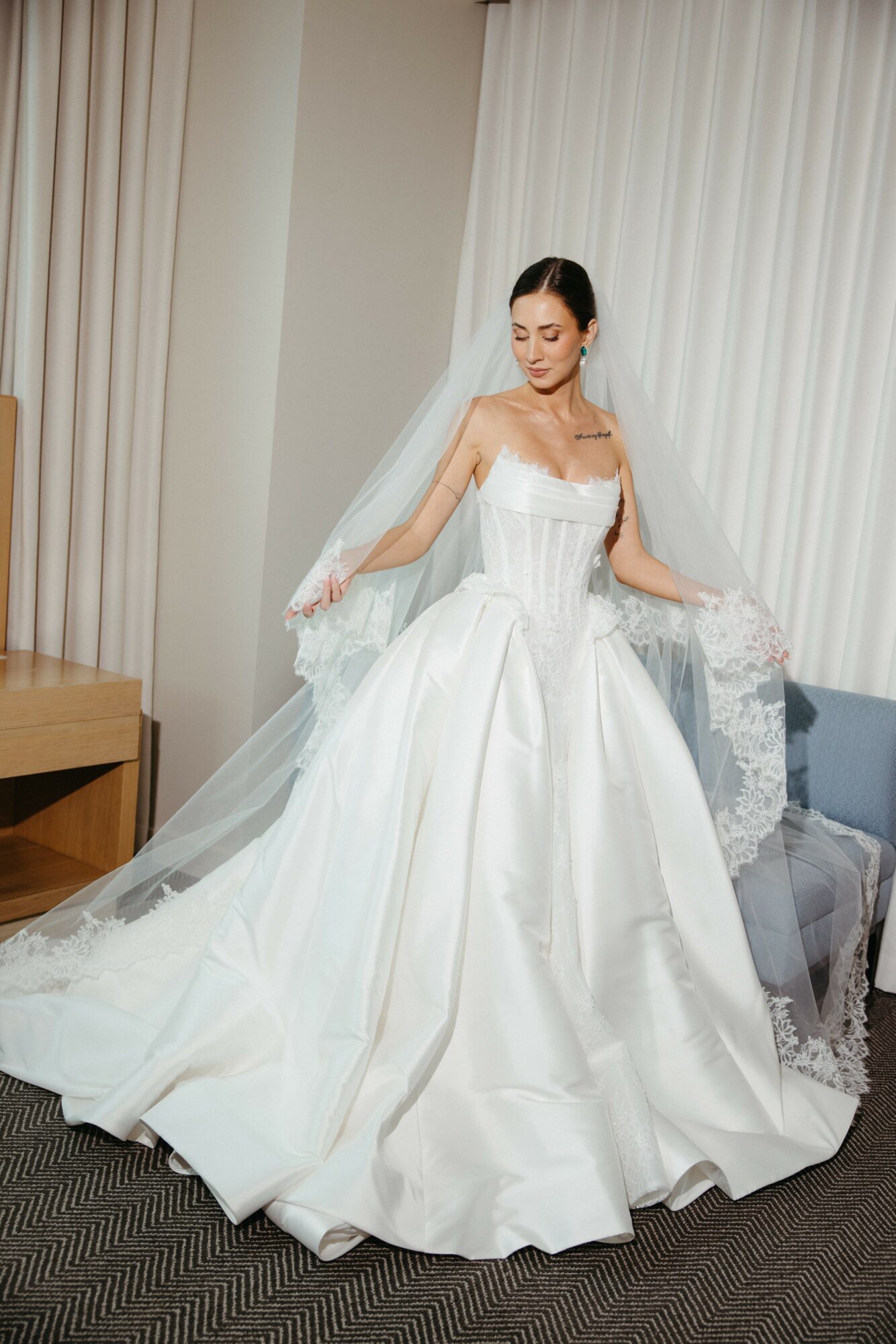 Woman in wedding dress standing indoors, holding the sides of her dress, with a veil and a long train.