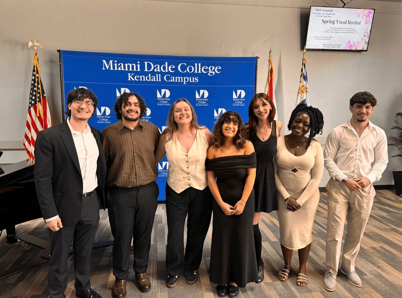 Group of seven diverse people standing in front of a Miami Dade College banner, smiling, indoors.