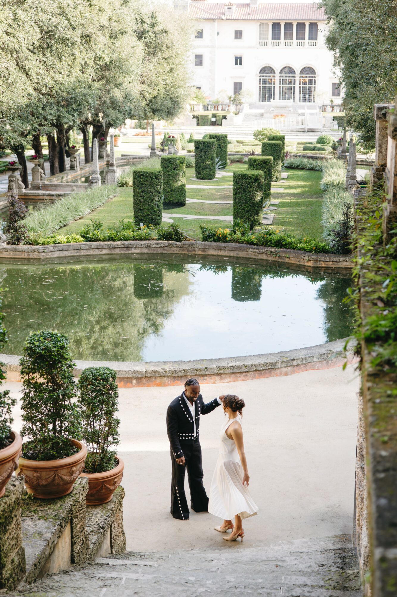 A couple stands near a pond in a garden with trimmed bushes and a large white building in the background.