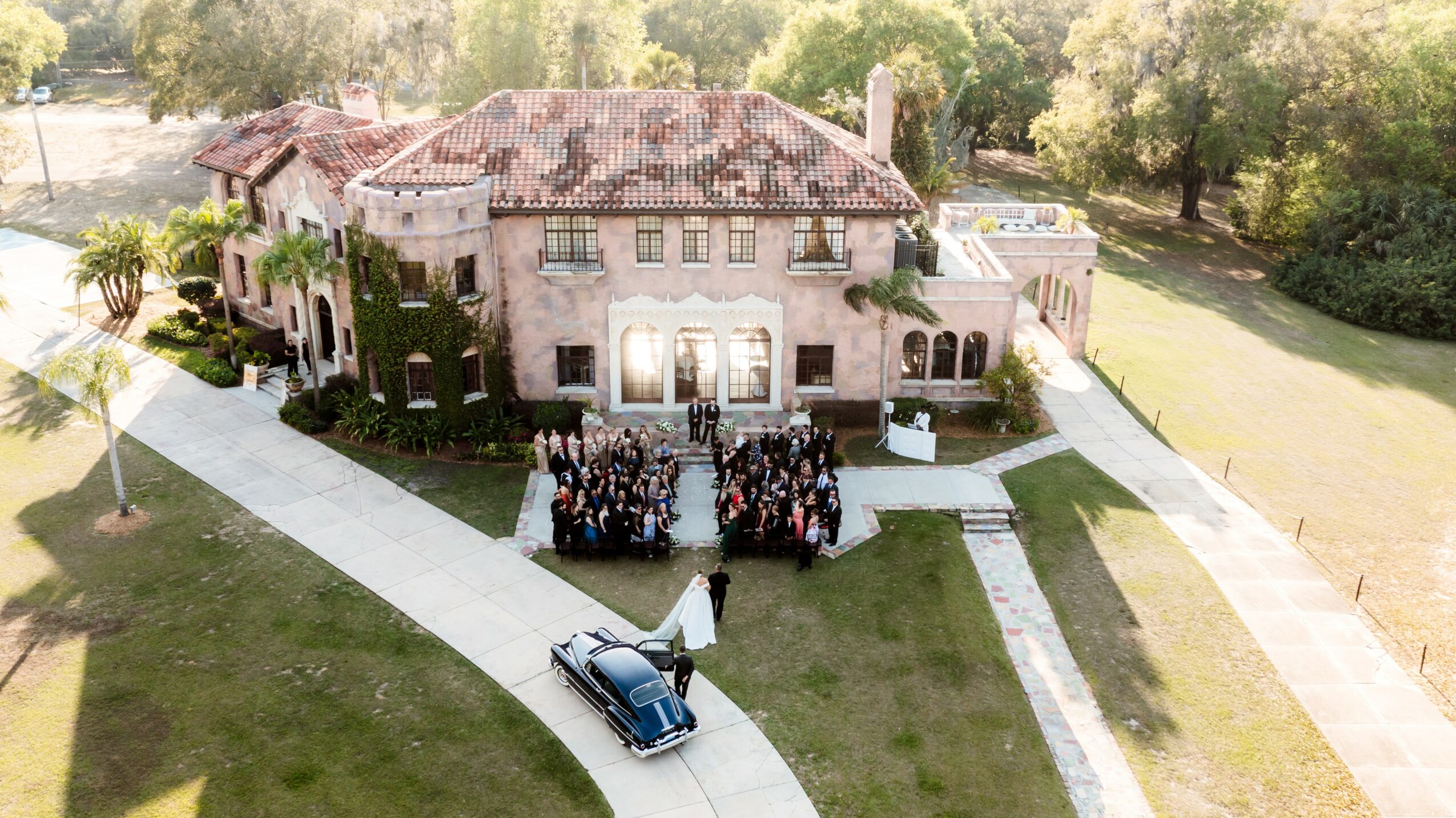 Large mansion with a group of people gathered in front, surrounded by trees and a driveway with a car.