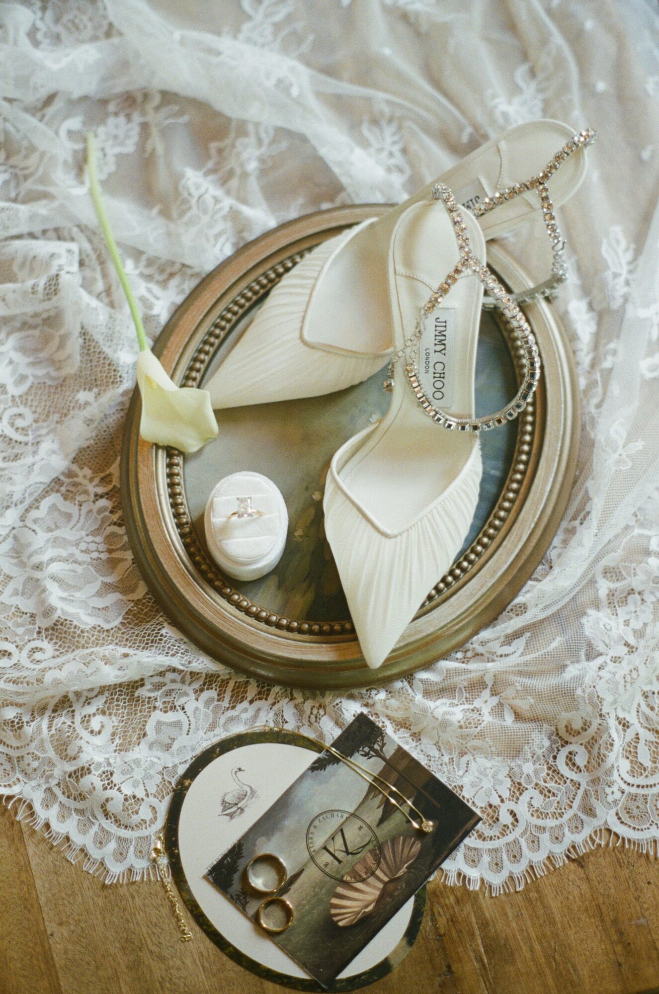 Cream-colored high-heeled shoes with ankle straps on a round mirror, lace fabric, and jewelry, including earrings and a ring.