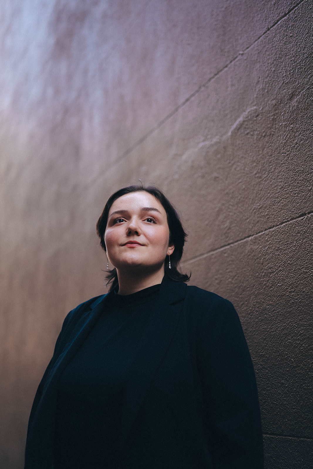 Woman with dark hair and black clothing standing against a textured wall, looking at the camera.