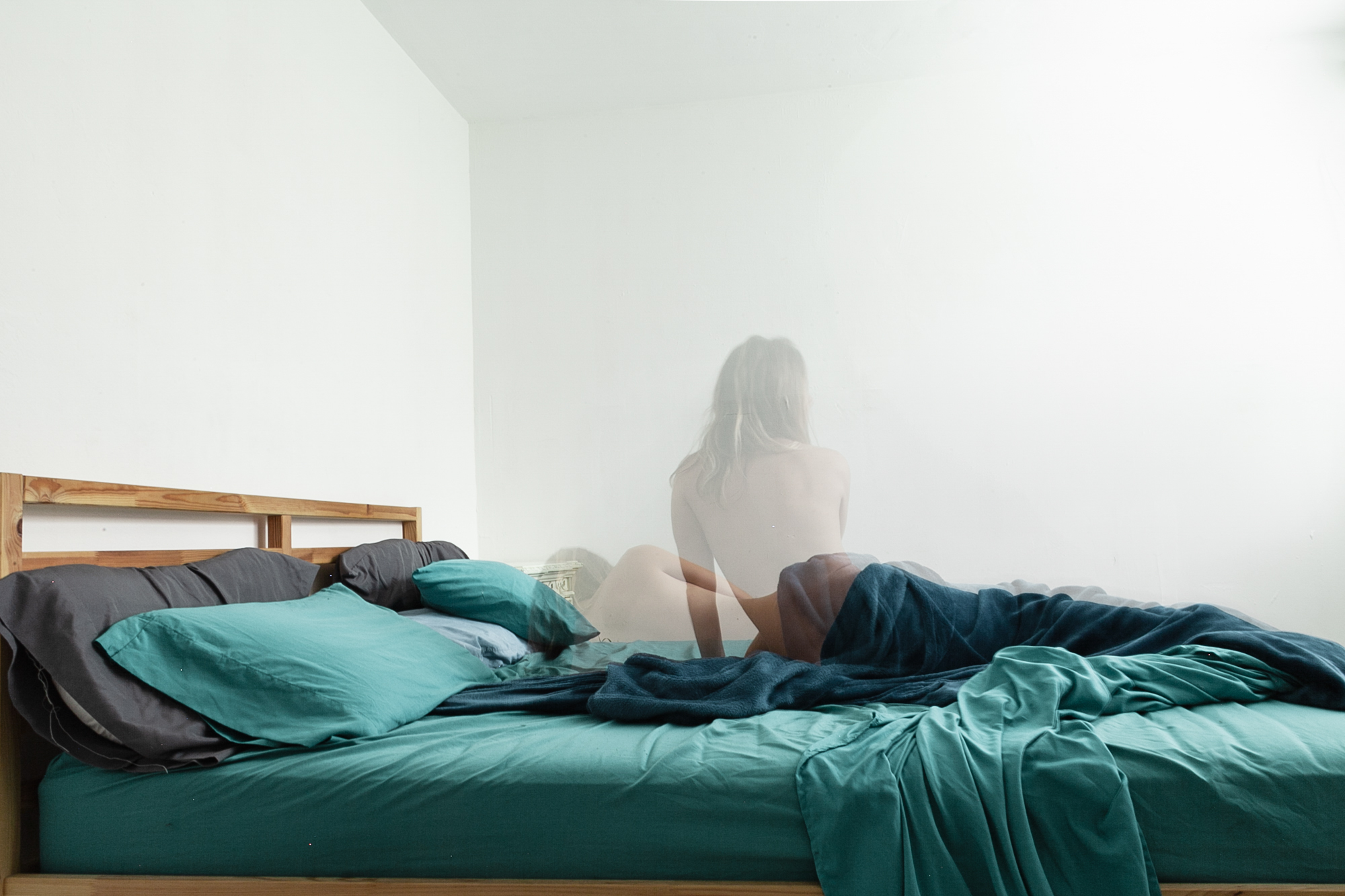 A person sitting on a bed with a foggy or semi-transparent overlay, in a room with white walls and a wooden headboard.