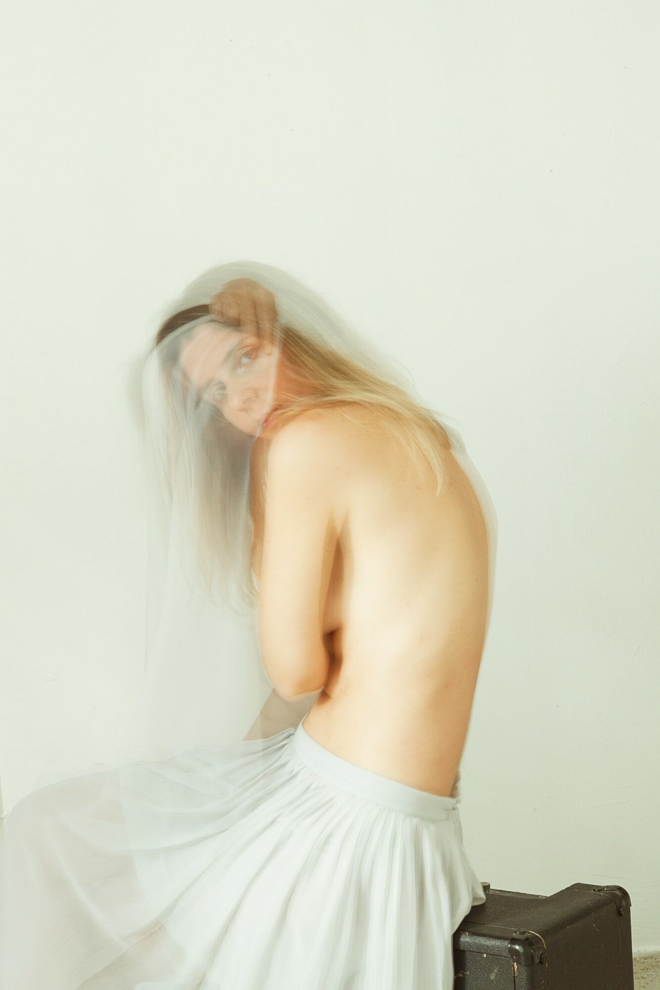 A person with long hair sits topless, wearing a white skirt, against a plain light background.